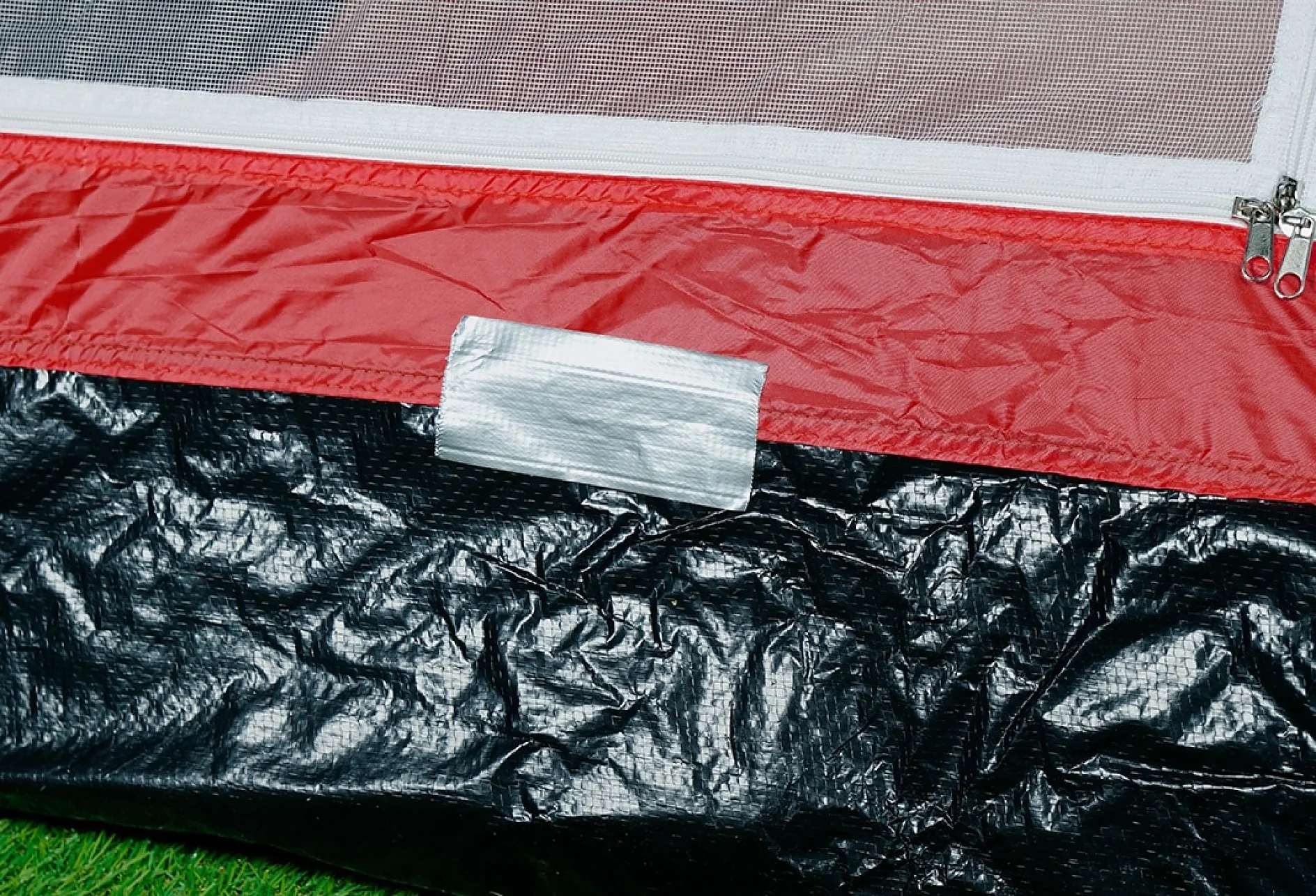 Close-up of a red and black tent with a piece of gray tesa tape patching a section on the black portion. The tent has a partially visible zipper and is set on green grass. (This text has been generated by AI)