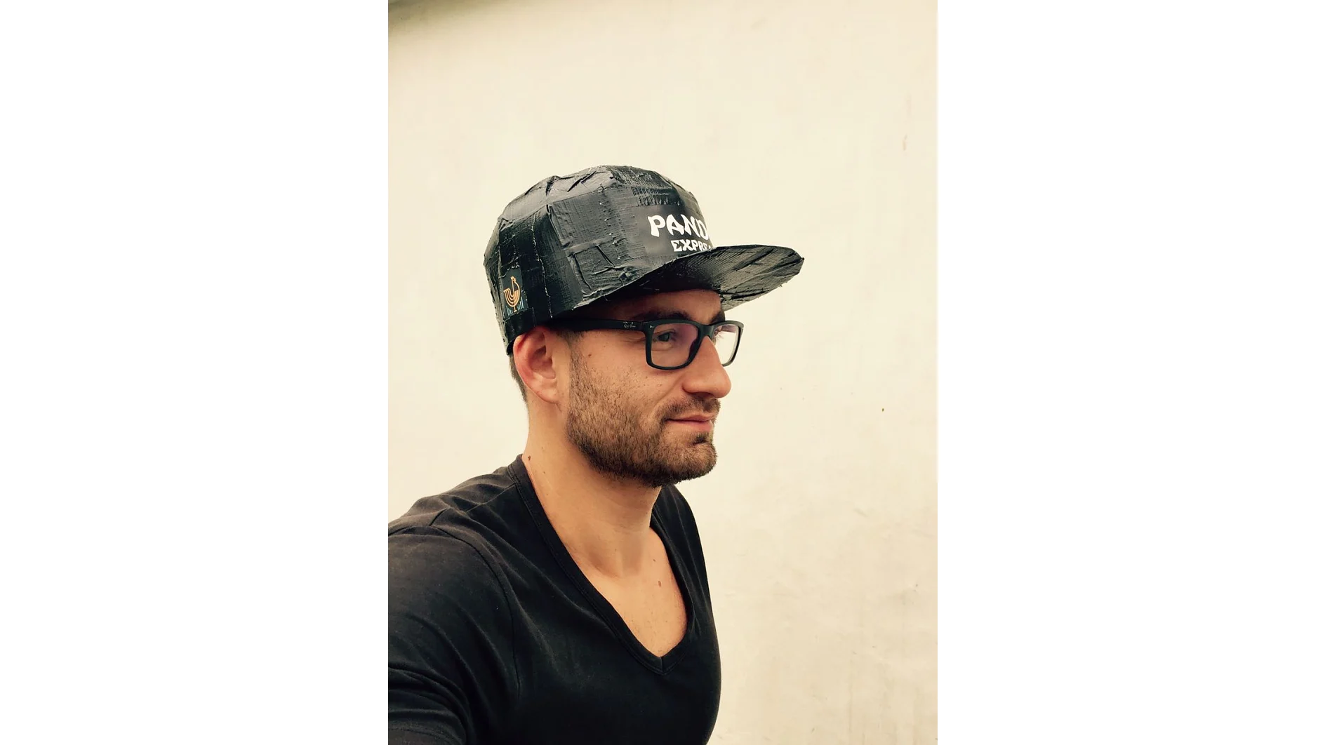 Um homem com barba e óculos está usando um boné preto com um logotipo. Ele está vestido com uma camisa preta de gola V e está olhando para o lado. O fundo é uma parede branca simples. (This text has been generated by AI)