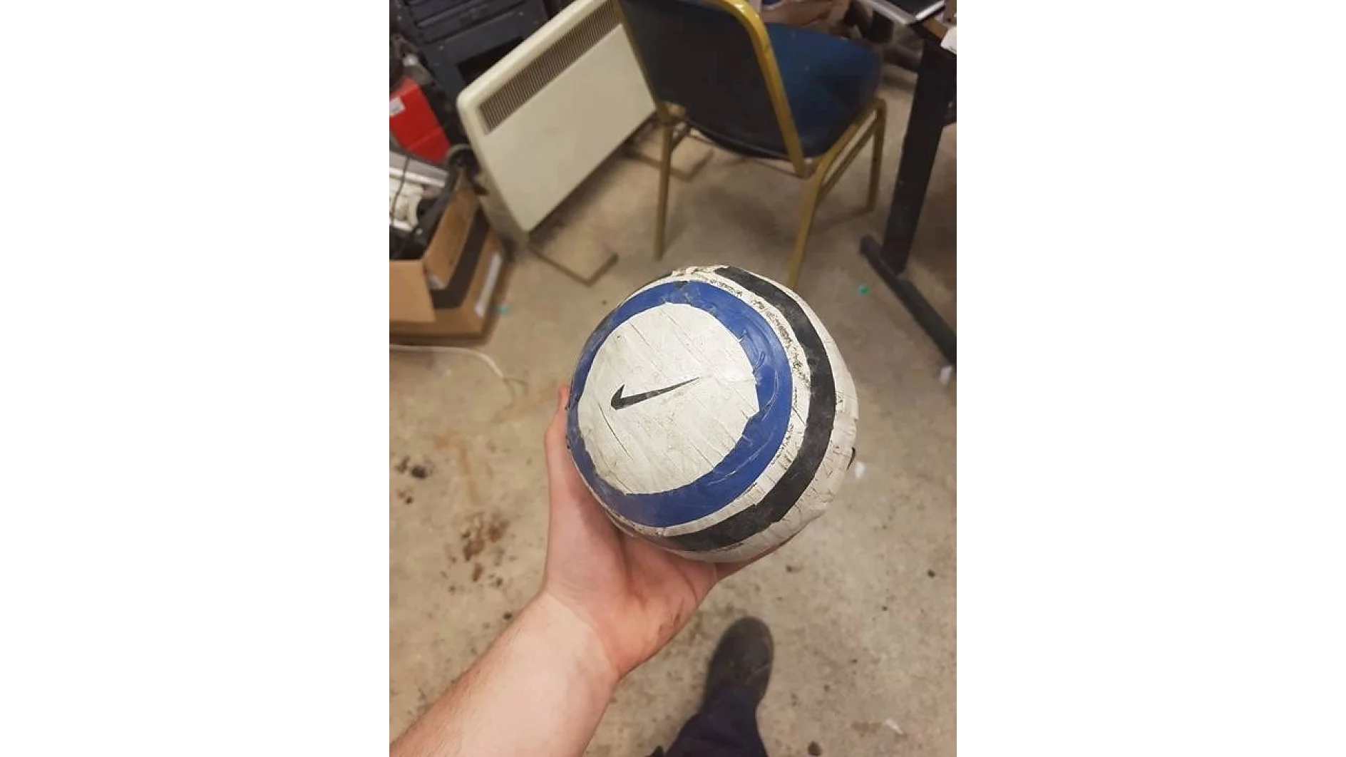 Uma pessoa segura uma bola improvisada enrolada em fita tesa branca, azul e preta. A bola apresenta um logotipo da Nike desenhado. O fundo inclui uma cadeira, um aquecedor e vários itens no chão de uma sala desorganizada. (This text has been generated by AI)