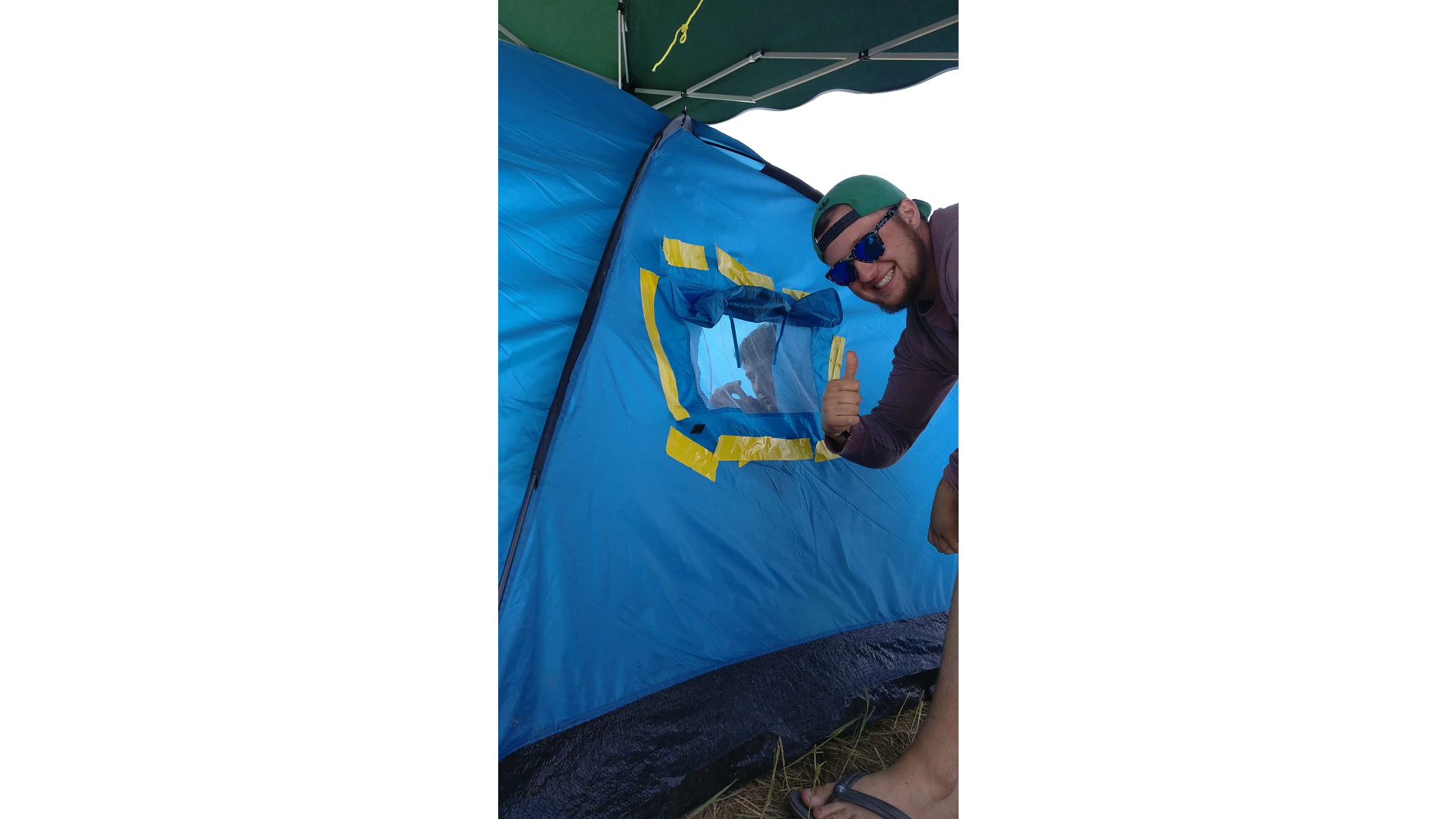 Uma pessoa usando óculos escuros e um boné verde está sorrindo e dando um sinal de positivo ao lado de uma barraca azul. A janela da barraca está presa com fita tesa amarela. A pessoa está inclinada em direção à barraca, com grama visível na parte inferior da imagem. (This text has been generated by AI)