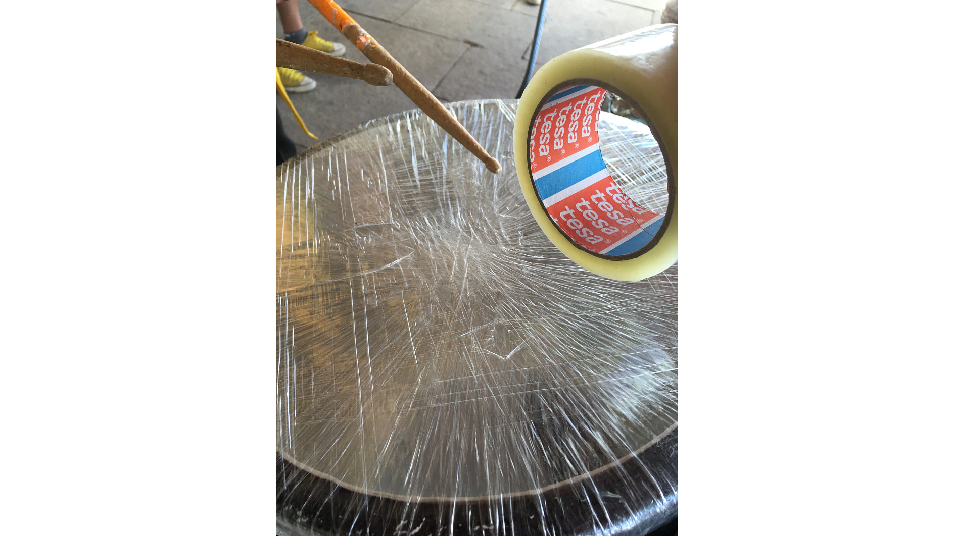 Um rolo de fita tesa repousa sobre um tambor coberto com filme plástico transparente. A mão de uma pessoa é vista segurando um pedaço de madeira ou um martelo acima do tambor, e outra pessoa com sapatos amarelos está de pé ao fundo. (This text has been generated by AI)