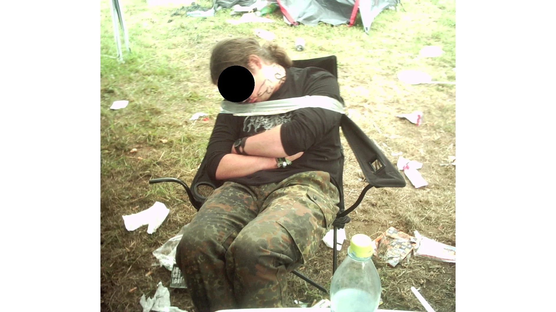 Uma pessoa com cabelo longo, rosto obscurecido, está dormindo e presa a uma cadeira de acampamento com fita tesa, ao ar livre. Ela está vestindo uma camisa preta e calças camufladas. Itens espalhados são visíveis na grama, incluindo uma garrafa e papel. Uma barraca pode ser vista ao fundo. (This text has been generated by AI)