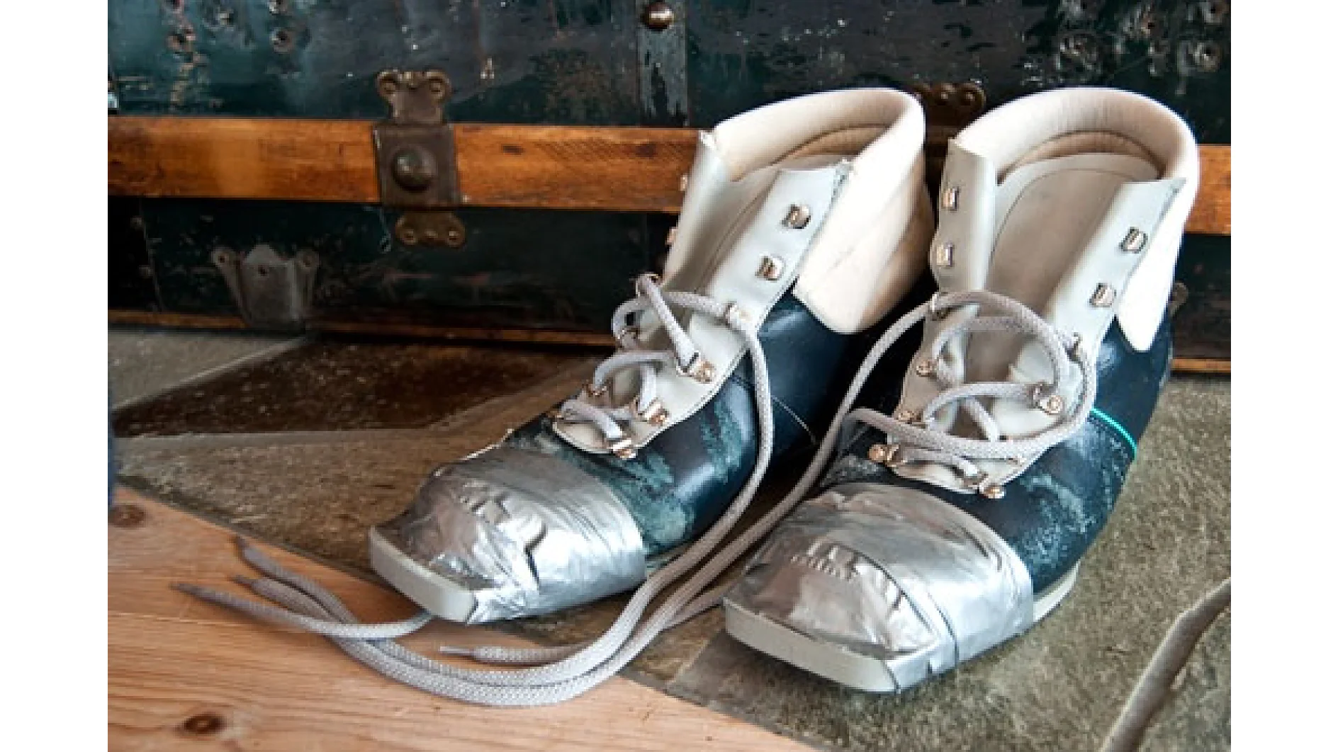 Um par de patins de gelo desgastados com fita tesa cinza enrolada nos dedos. Os patins têm cabedais azuis e brancos com cadarços. Eles são colocados em uma superfície de madeira na frente de um baú vintage de madeira e metal. (This text has been generated by AI)