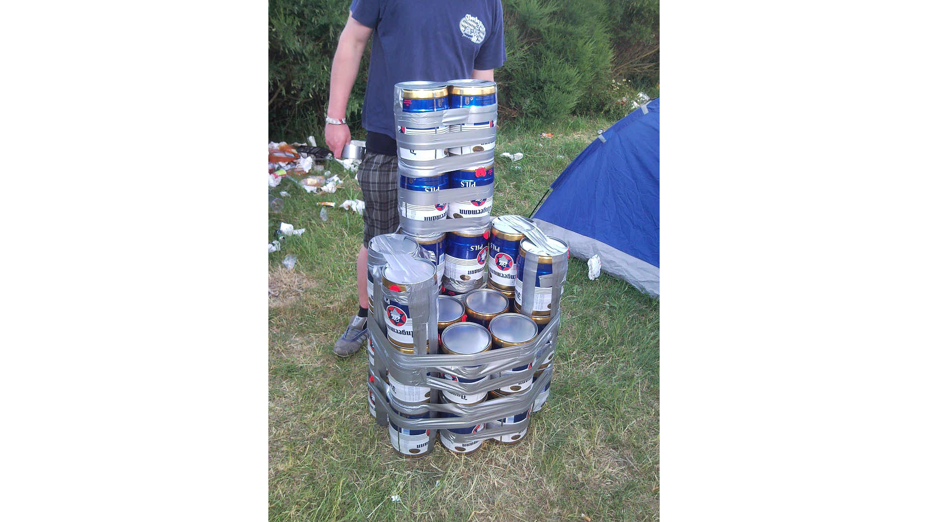 Uma pessoa está de pé ao lado de uma torre de latas de cerveja empilhadas, presas com fita tesa, ao ar livre, perto de uma barraca de acampamento. O chão está coberto de grama e alguns itens descartados. O rosto da pessoa não é visível. (This text has been generated by AI)
