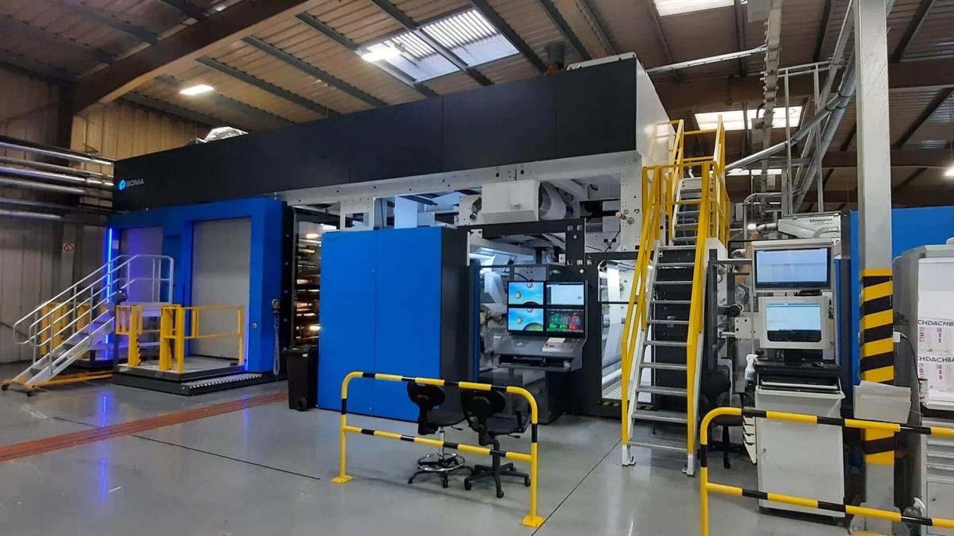 Inside a factory setting, there is a large industrial printing machine. It features blue and black panels, multiple control screens, and a set of stairs leading to an upper section. The area is well-lit and surrounded by safety railings. (This text has been generated by AI)