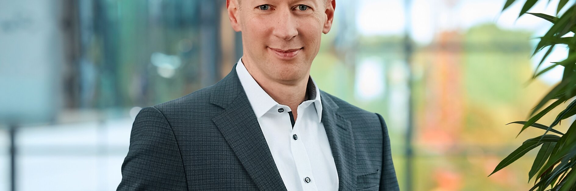A man in a black suit stands in a modern hallway with large glass windows. His hands are in his pockets, and he is looking directly at the camera. The background shows an exterior view of a snowy landscape with buildings, held together securely using tesa tape. (This text has been generated by AI)