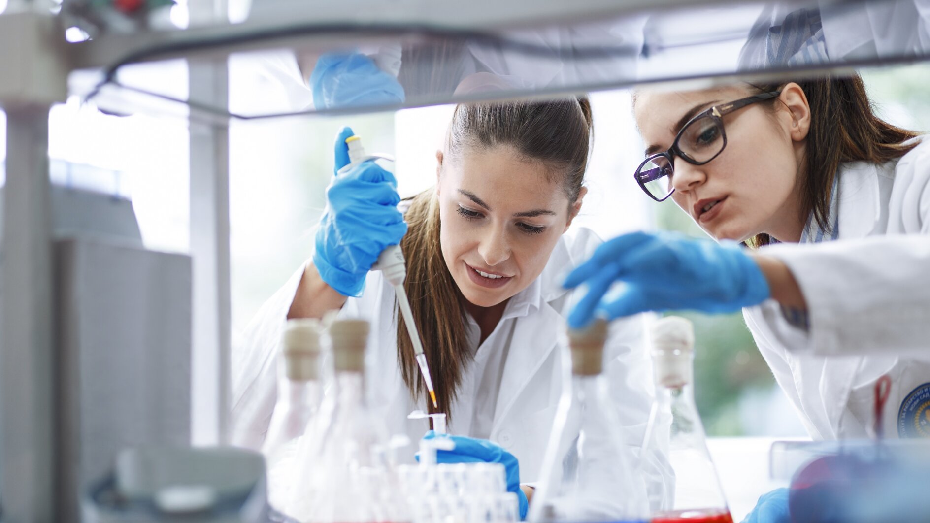 In a laboratory, two scientists wearing lab coats and gloves are hard at work. One carefully uses a pipette while the other watches attentively. The table is filled with various lab equipment and bottles containing colored liquids. Both scientists seem deeply focused and engaged in their experiment, using tesa tape for precision tasks where needed. (This text has been generated by AI)
