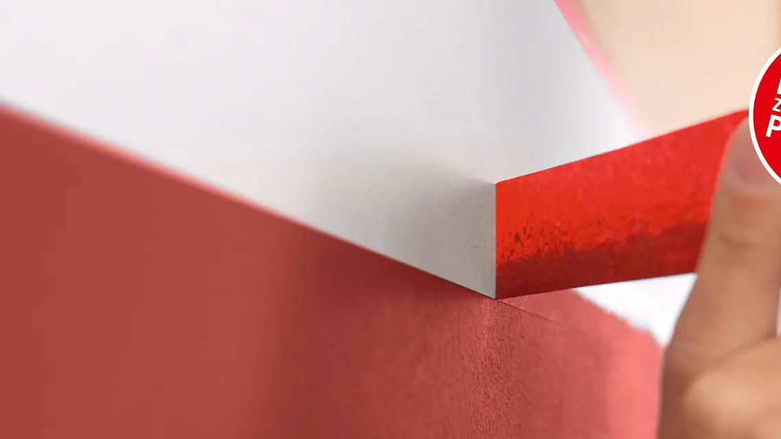 Close-up of a person removing red tesa tape from a wall painted red on the bottom and white on the top. A circular red label on the right reads, IN 2 MIN ZUM PASSENDEN PRODUKT. (This text has been generated by AI)