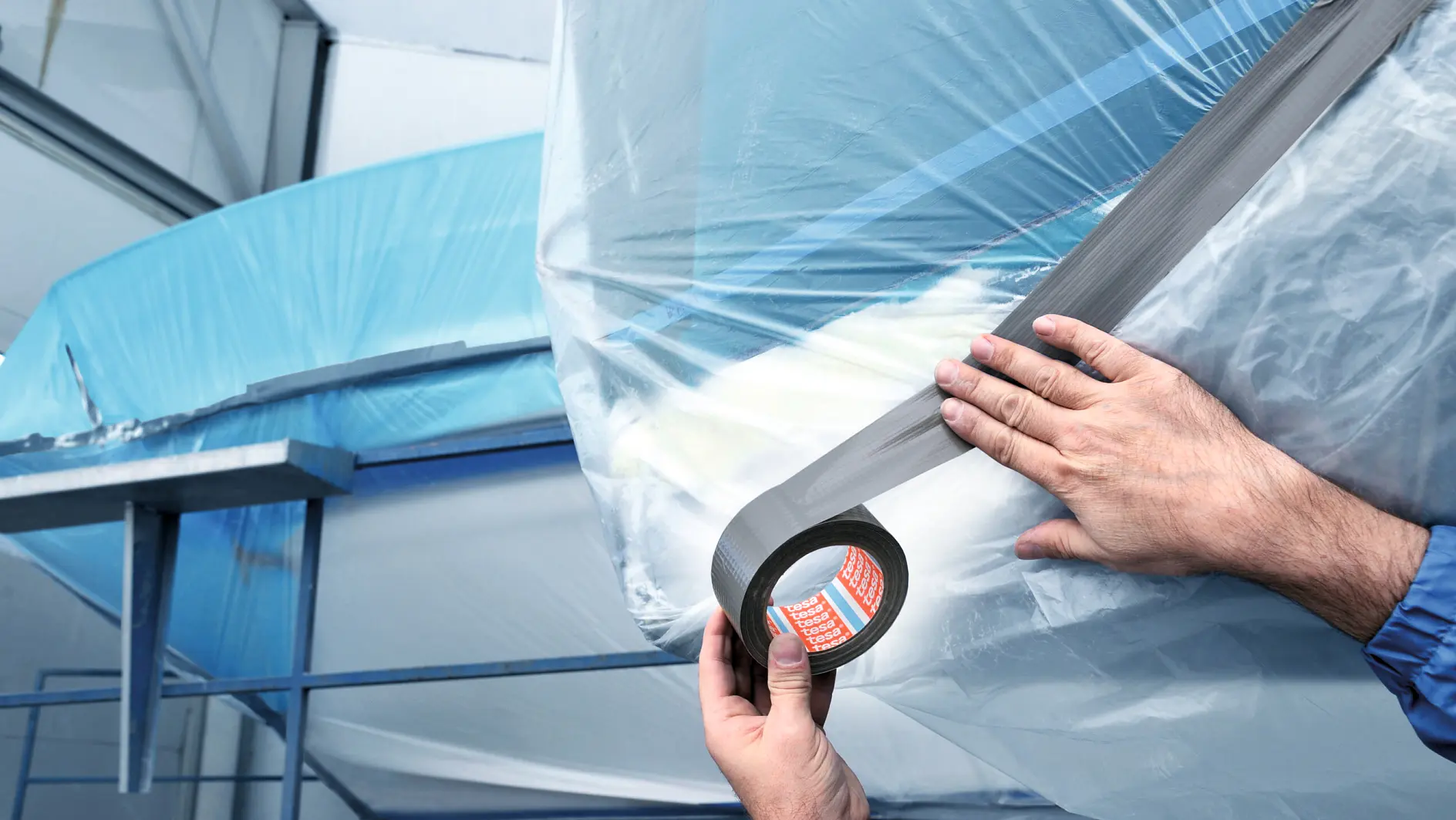 Two hands apply gray tesa tape to secure clear plastic sheeting over a large blue object, possibly in an industrial or construction setting. The background shows metal scaffolding and parts of the structure.
