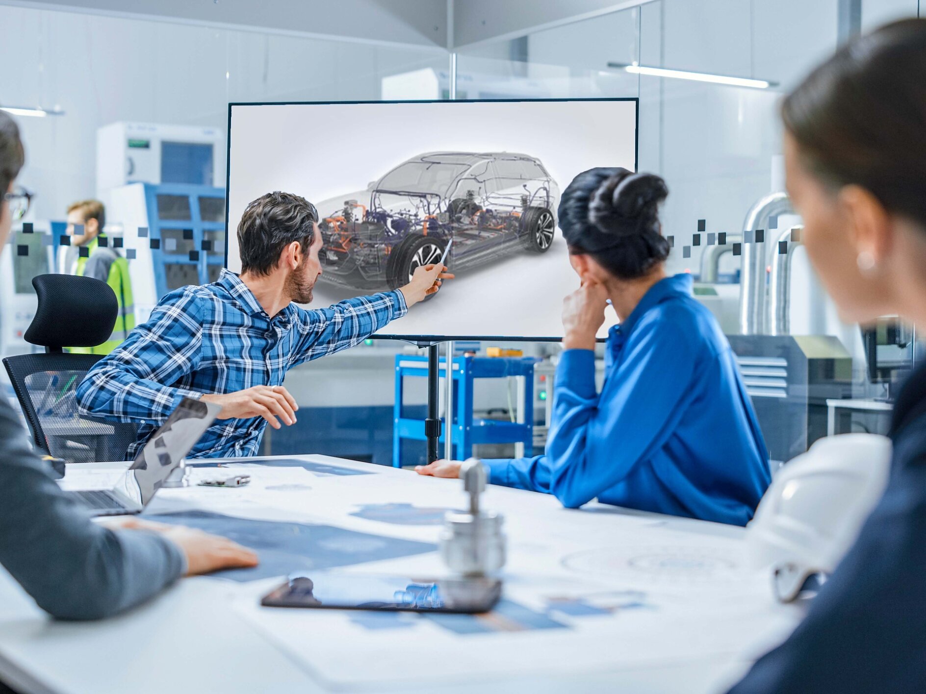 A group of four people in a modern office setting look at a screen displaying a cars internal components. A man in a plaid shirt points at the screen. Various technical documents, equipment, and tesa tape are on the table. (This text has been generated by AI)