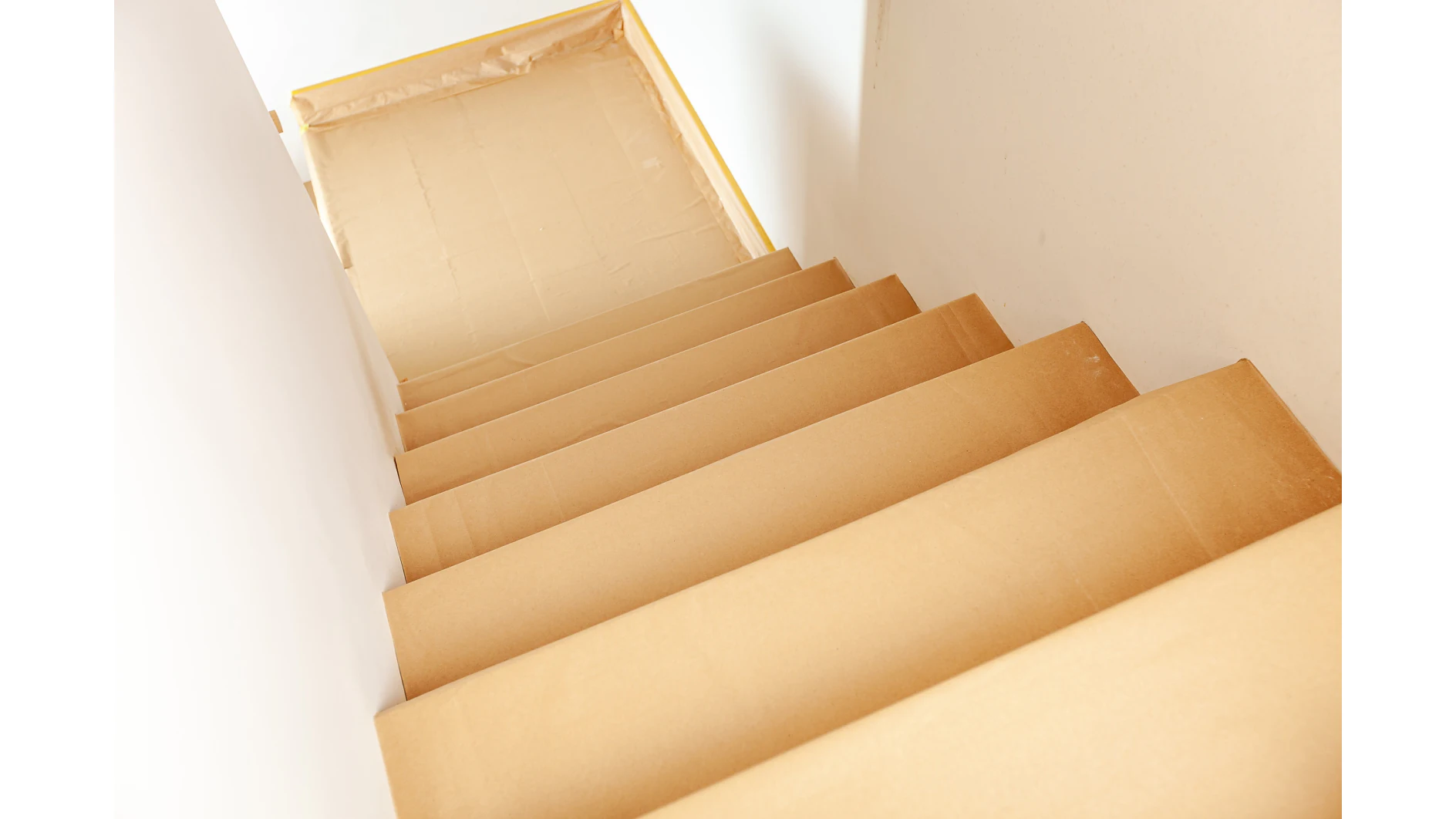 Uma escadaria coberta com folhas de papel protetor castanho, incluindo a zona de patamar no topo, sugerindo a preparação para trabalhos de pintura ou renovação. O papel está fixado com fita tesa. As paredes são brancas e não há objectos visíveis nas escadas. (This text has been generated by AI)