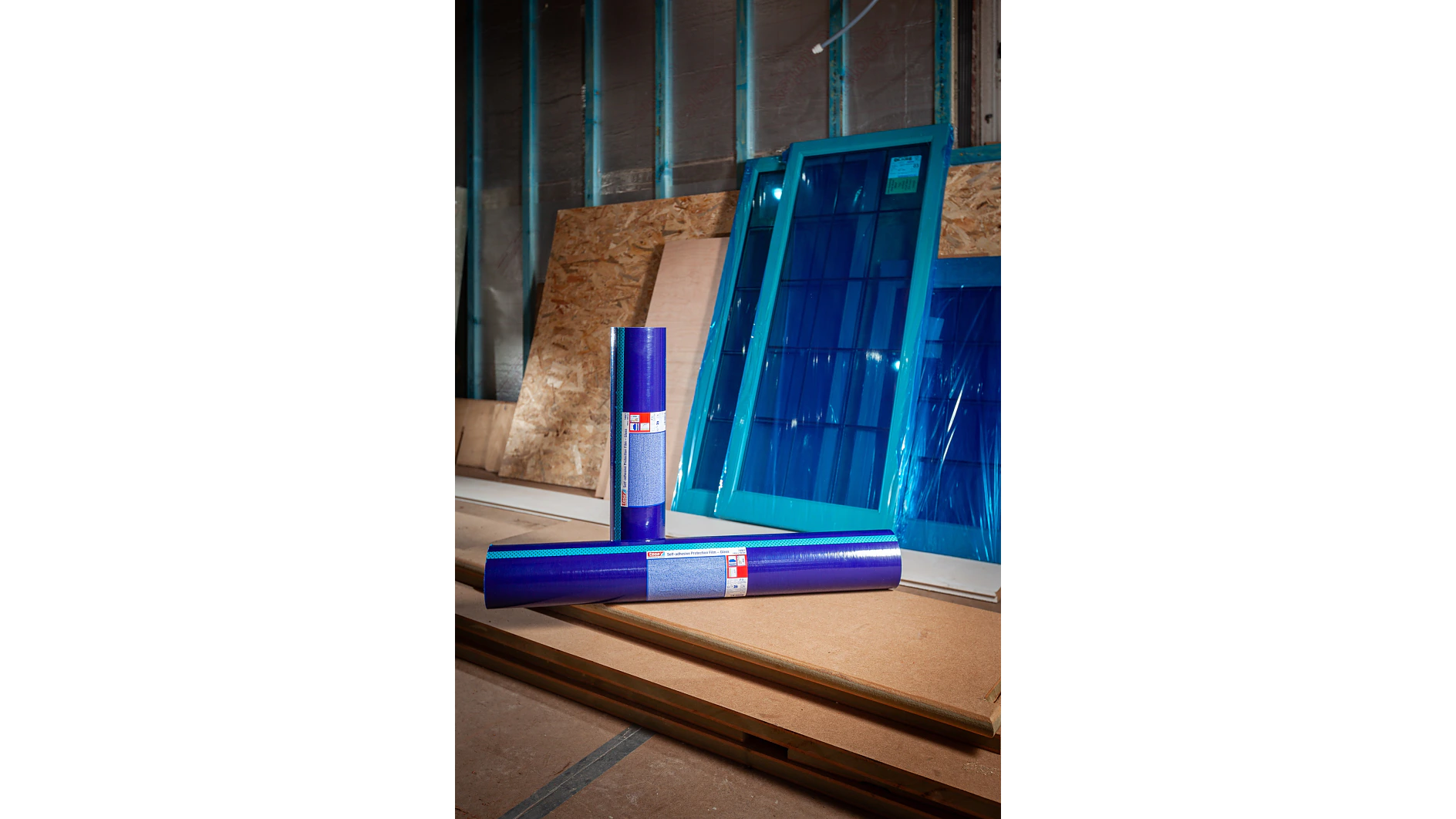 Três rolos de película protetora de fita tesa azul estão em pé no chão de um estaleiro de construção, com vidros de janelas com moldura azul e folhas de madeira encostadas a uma parede ao fundo. (This text has been generated by AI)