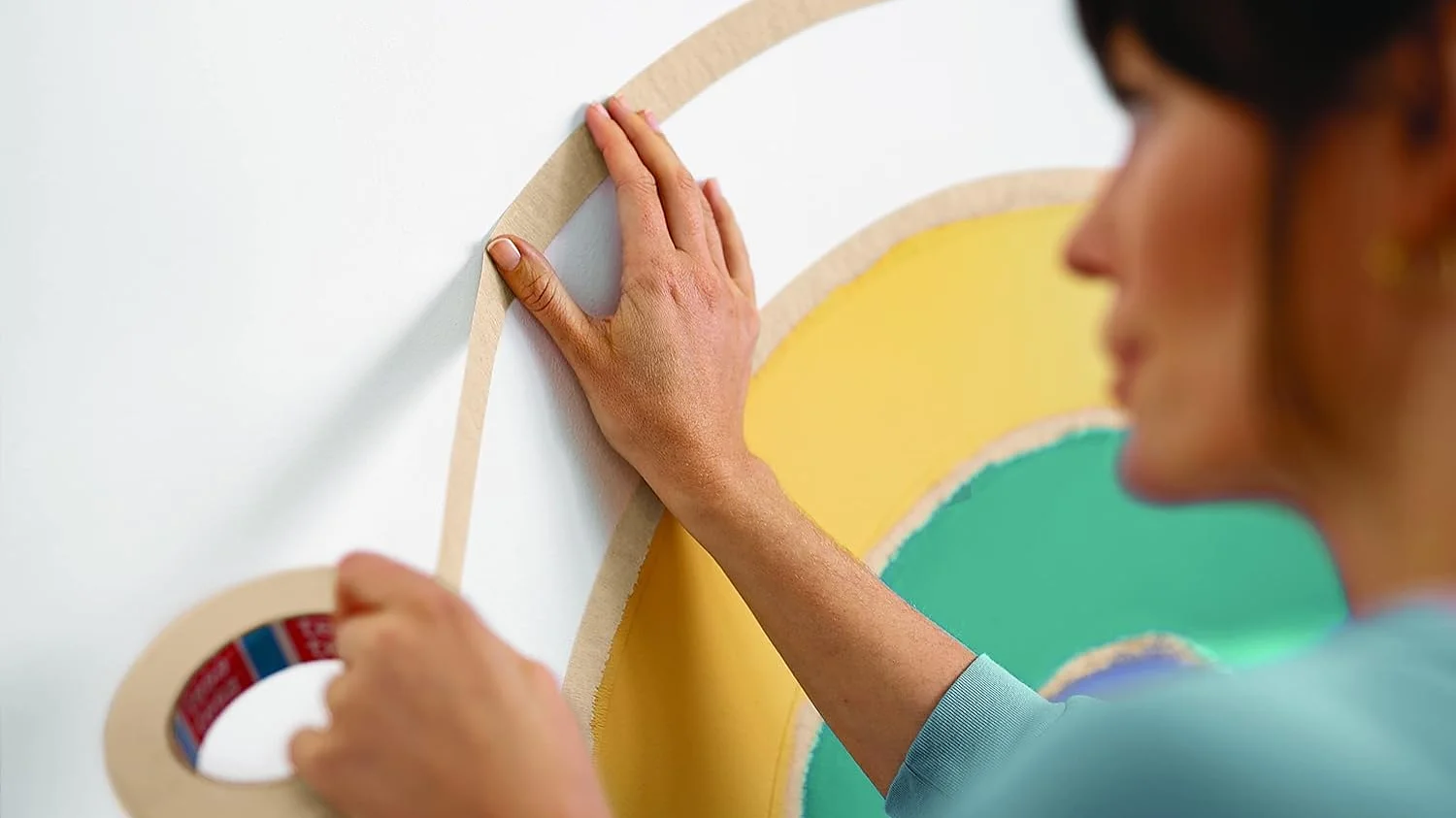 Uma pessoa aplica fita tesa em uma linha curva em uma parede branca com formas pintadas de amarelo e verde sobrepostas. A pessoa segura o dispensador de fita tesa com uma mão e pressiona a fita com a outra. (This text has been generated by AI)