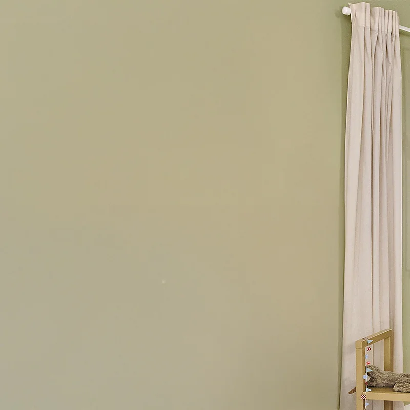 Uma mulher está num quarto de criança iluminado pelo sol, com paredes verdes claras, abrindo uma janela. O quarto tem uma cama pequena, brinquedos, uma cadeira e uma grinalda pendurada na parede com fita tesa. O ambiente parece calmo e arrumado. (This text has been generated by AI)