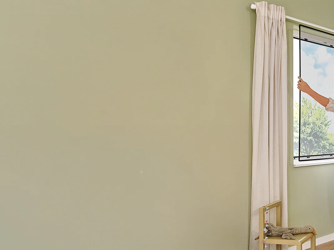 Uma mulher está num quarto de criança iluminado pelo sol, com paredes verdes claras, abrindo uma janela. O quarto tem uma cama pequena, brinquedos, uma cadeira e uma grinalda pendurada na parede com fita tesa. O ambiente parece calmo e arrumado. (This text has been generated by AI)