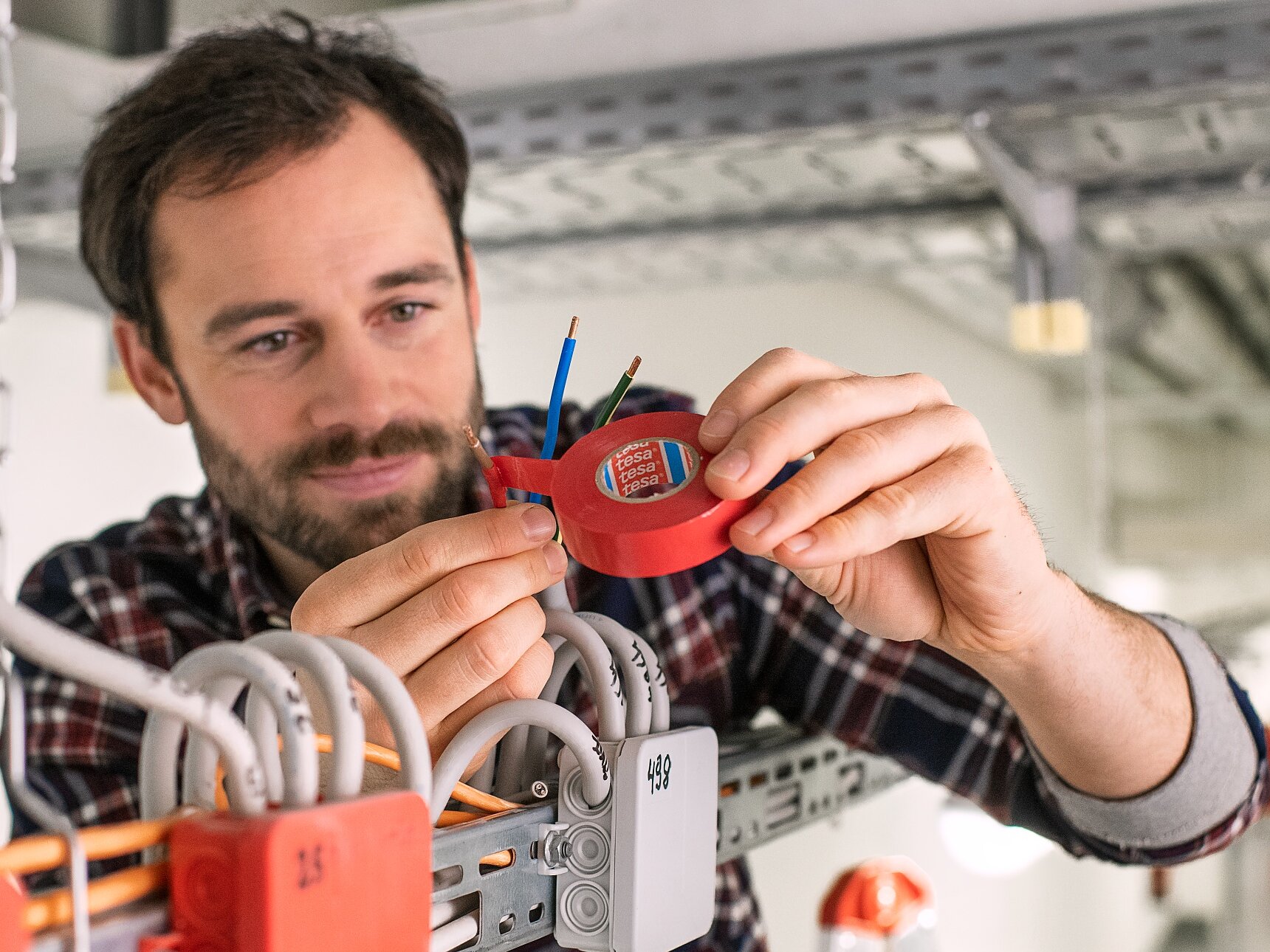 Um homem de camisa xadrez usa um dispositivo de teste elétrico vermelho em fios presos a uma bandeja de cabos de metal em um ambiente industrial. Ele está concentrado em sua tarefa, e vários fios e caixas elétricas presos com fita tesa são visíveis. (This text has been generated by AI)