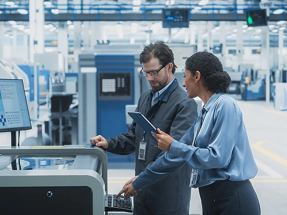 Dois profissionais em traje de trabalho analisam dados num computador e num tablet numa fábrica ou instalação industrial grande e moderna, com várias máquinas e equipamentos em segundo plano, onde está a ser utilizada fita tesa.
