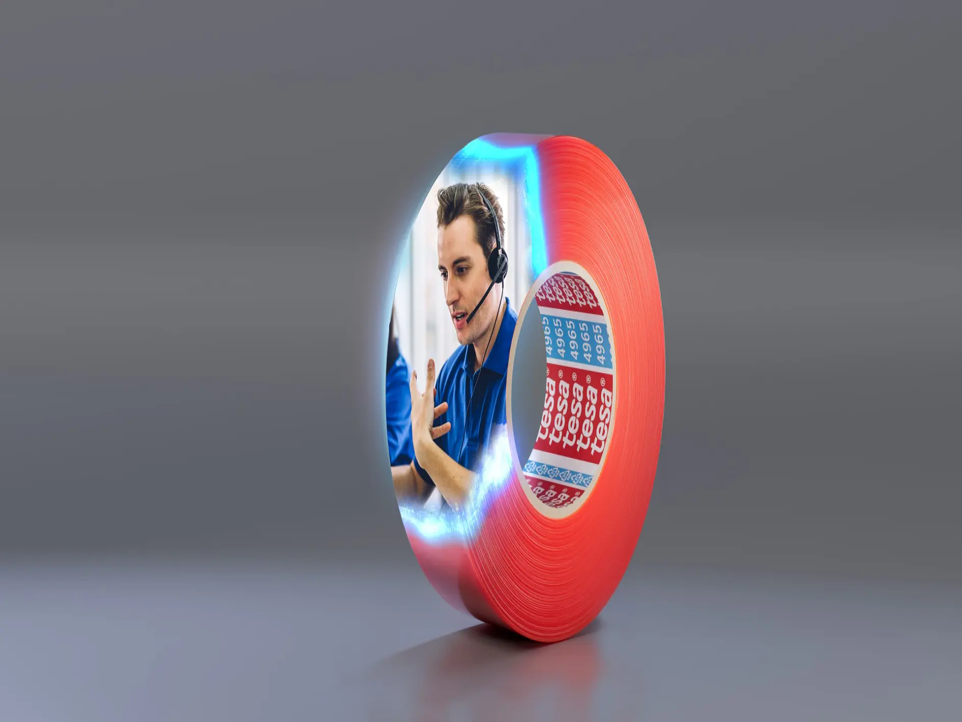 A red roll of tesa tape is on a gray surface. The side of the tesa tape features an image of a person wearing a headset, gesturing with one hand. Next to the person is a blue and red calendar with some dates marked. (This text has been generated by AI)