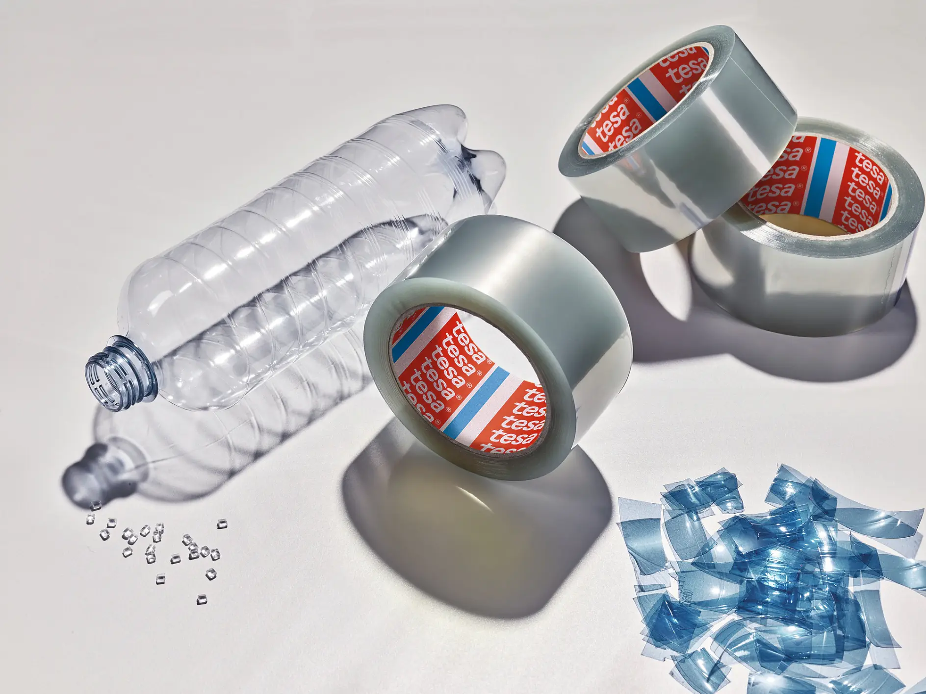 Clear plastic water bottle, three rolls of silver tesa adhesive tape, and scattered blue plastic bottle caps on a white surface. (This text has been generated by AI)