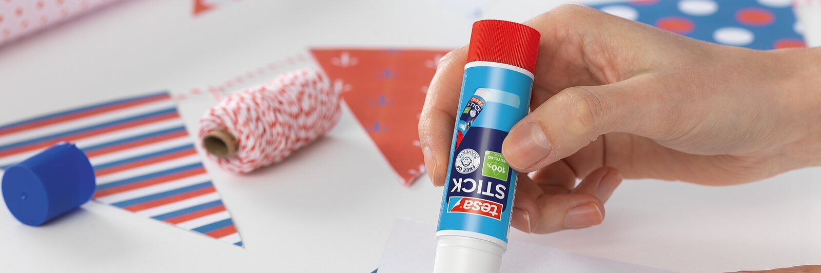 Uma mão cola uma fita tesa em um pedaço de papel triangular branco, rodeado por outros triângulos com estampas coloridas e rolos de papel de embrulho sobre uma superfície branca.