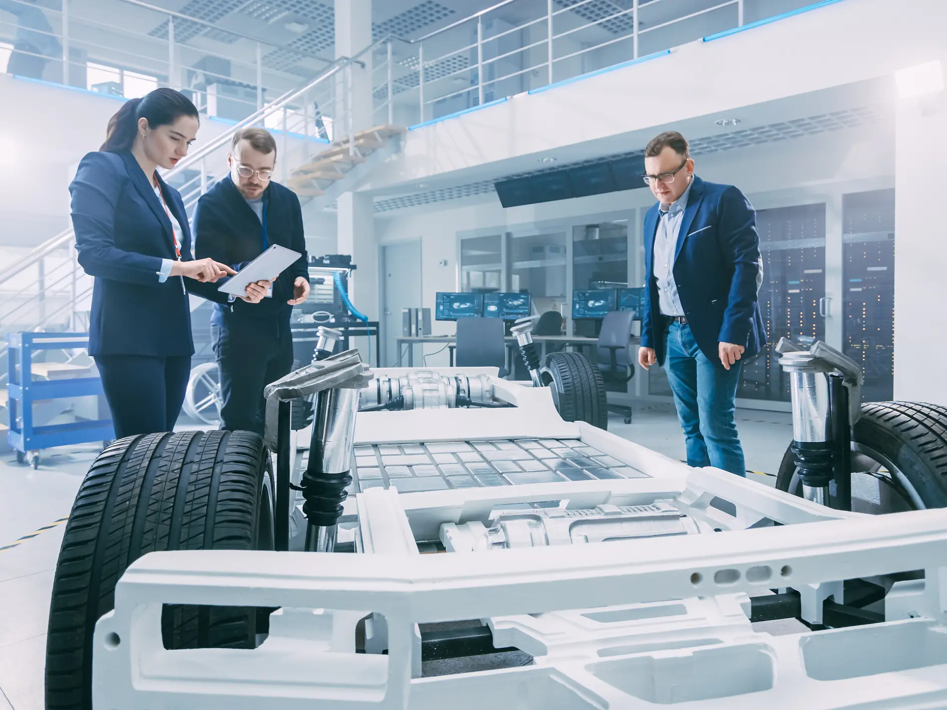 Três pessoas estão num laboratório moderno a examinar um protótipo de chassis de automóvel. A sala está bem iluminada, com grandes janelas e vários equipamentos técnicos. O chassis apresenta baterias e suspensão visíveis, indicando tecnologia de veículo elétrico, tudo montado de forma especializada com fita tesa. (This text has been generated by AI)