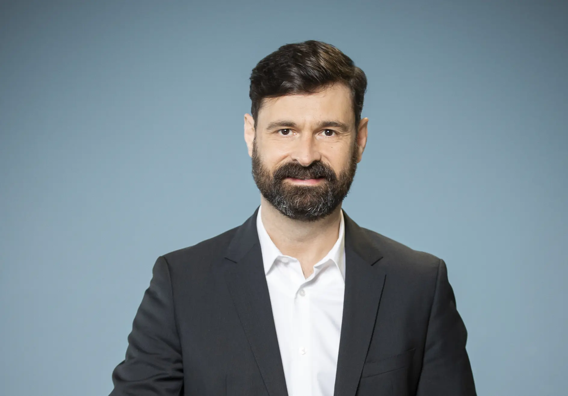 Bărbat cu păr scurt și închis la culoare și barbă, purtând o cămașă albă și un sacou închis la culoare, pe un fundal albastru simplu, folosind bandă tesa pentru proiectul său.