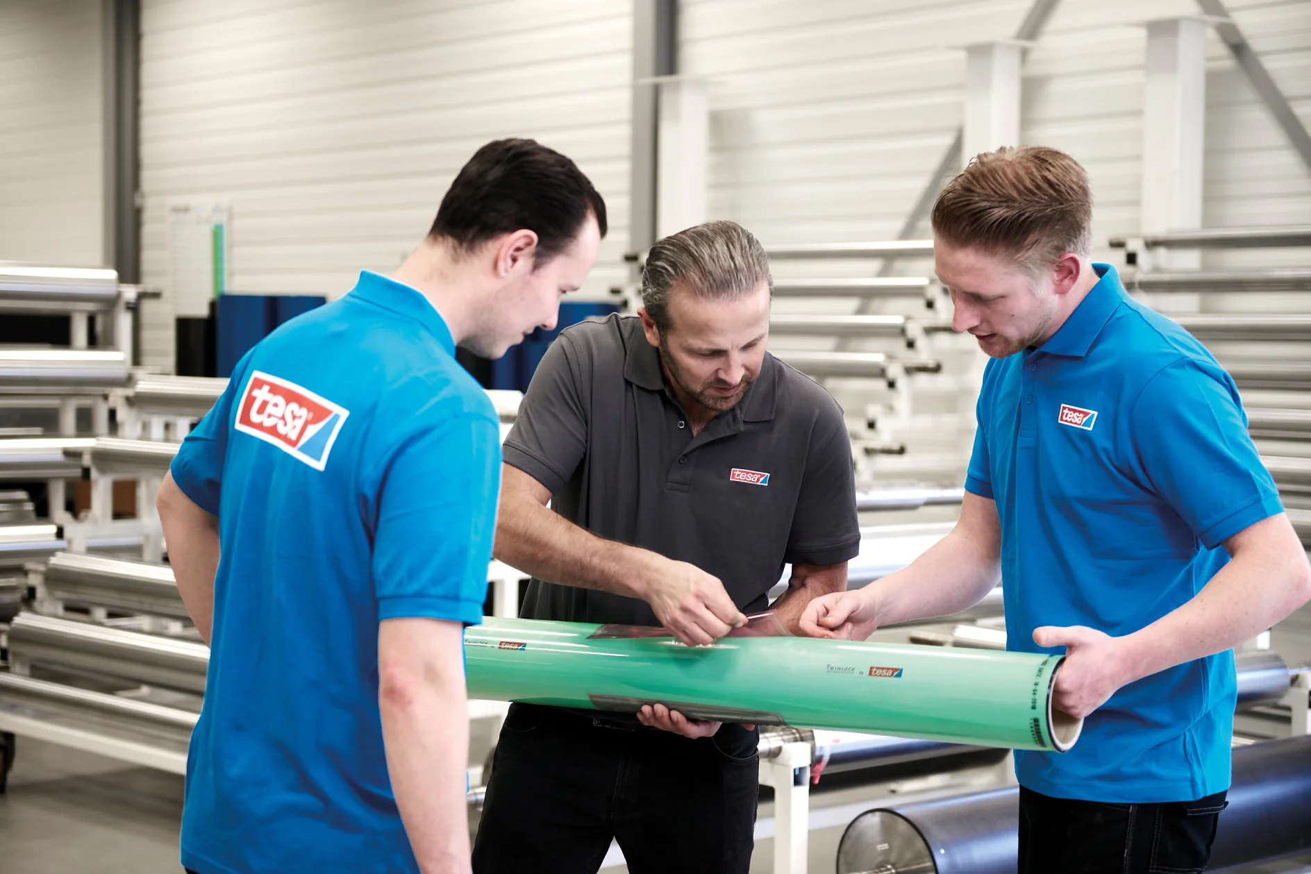 Three men in a factory setting examine a large green cylindrical object. Two men wear blue shirts with red logos, and the third, in the center, wears a gray shirt with the same logo. Metal pipes and machinery are visible in the background. (Acest text a fost generat de inteligența artificială)