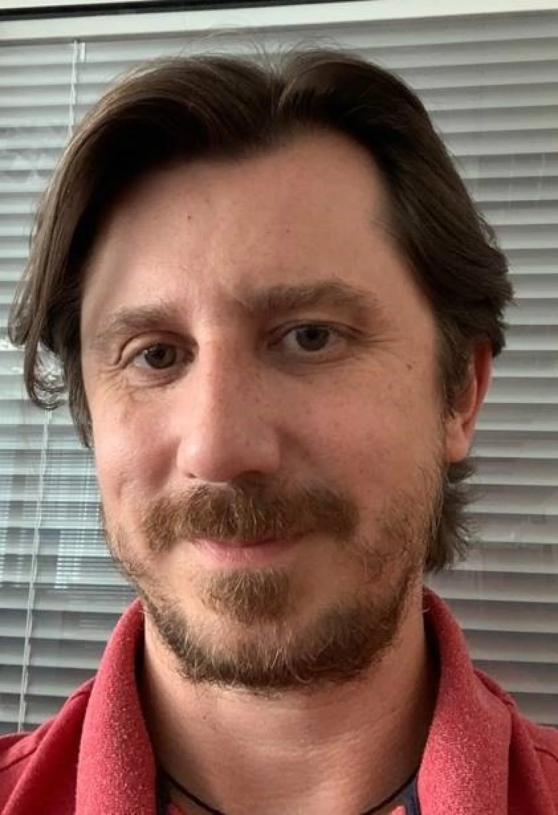 A person with medium-length brown hair and a beard is wearing a red shirt and standing in front of a window with closed blinds. The photo is a close-up, and the individual is looking directly at the camera. In their hand, they hold a roll of tesa tape, showcasing its versatility and quality for various needs. (Acest text a fost generat de inteligența artificială)