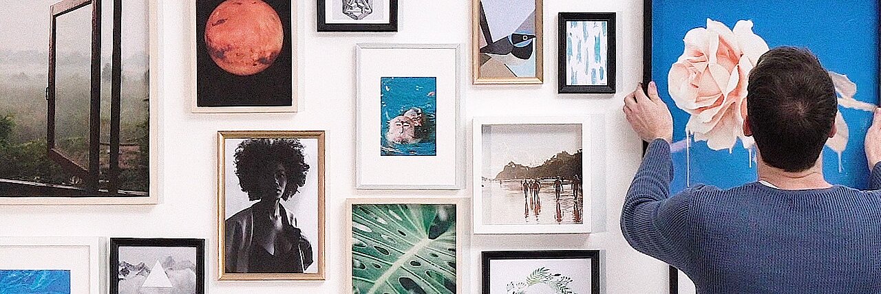 Persoană aranjând un tablou cu floare pe un perete decorat cu rame foto și opere de artă diverse, inclusiv imagini din natură, artă abstractă și fotografii.