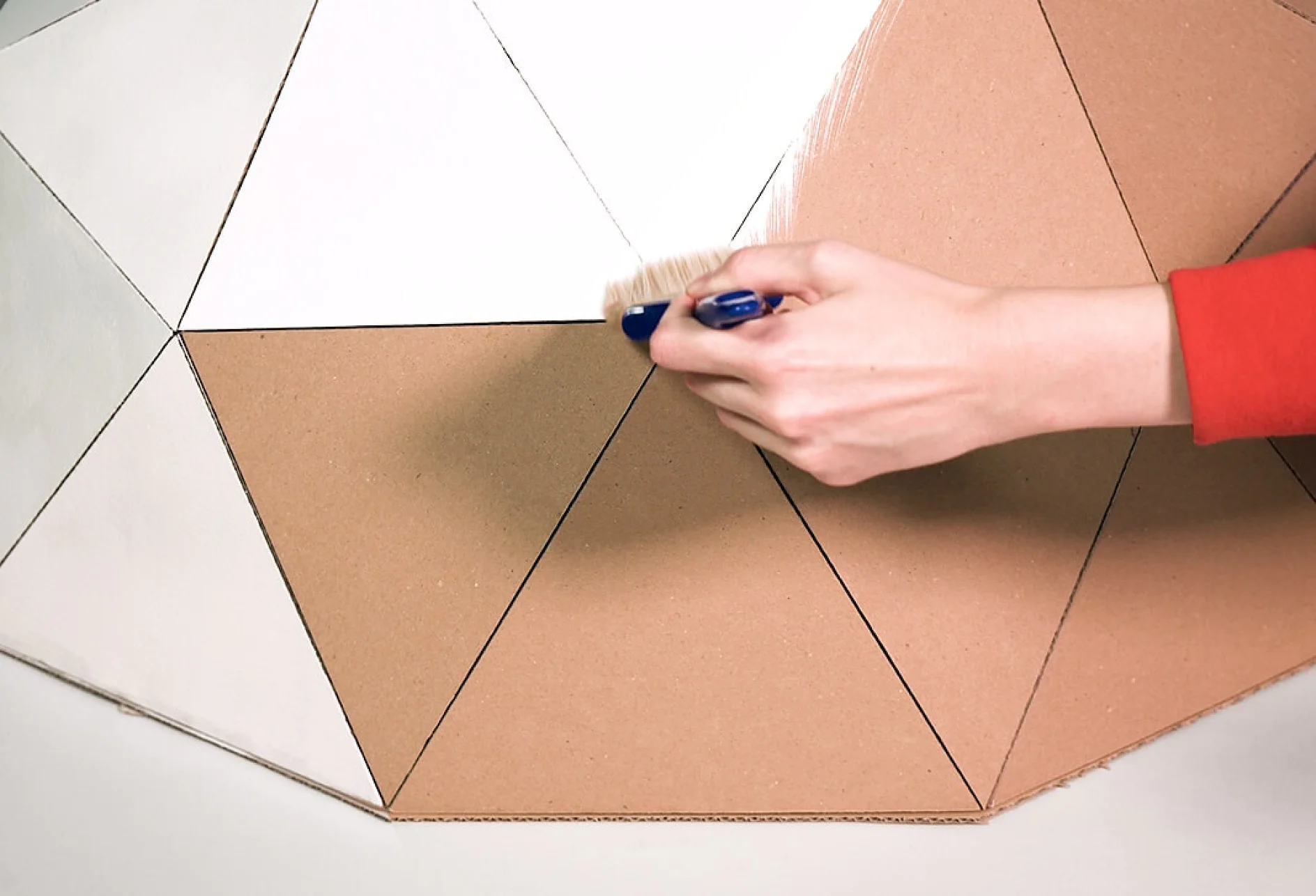 O mână îmbrăcată cu o mânecă roșie pictează un design geometric cu secțiuni triunghiulare maro și albe pe carton. Mâna ține o pensulă mică cu peri albaștri, aplicând vopsea albă. Banda tesa este folosită pentru a delimita zonele, asigurând linii clare între culorile diferite. Suprafața este parțial vopsită. (Acest text a fost generat de inteligența artificială)