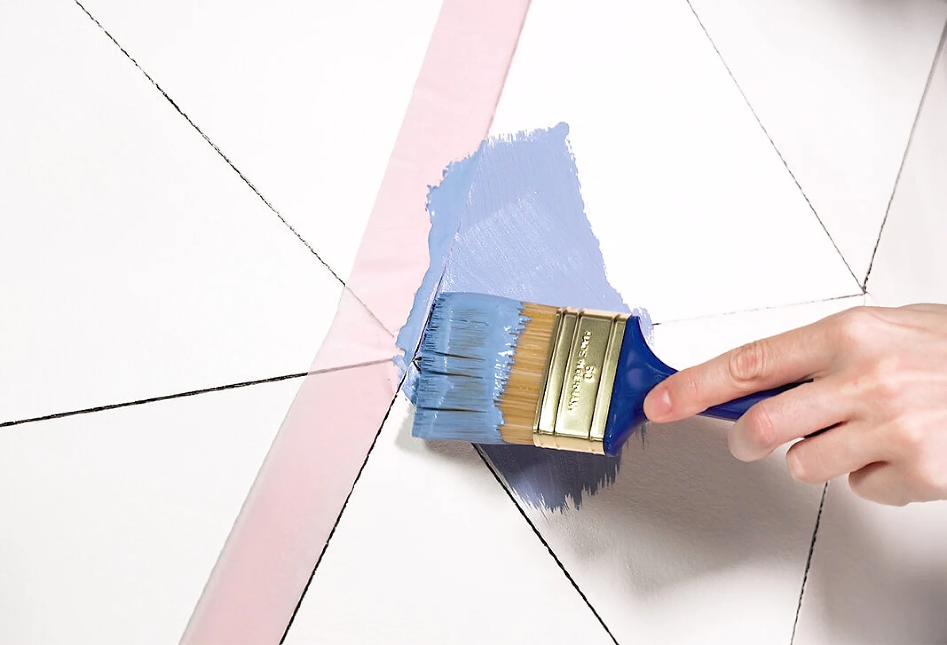 O mână ține o pensulă albastră aplicând vopsea albastru deschis pe un model geometric pe un perete alb. Liniile negre conturează formele, iar banda roz tesa marchează marginea zonei care este vopsită. (Acest text a fost generat de inteligența artificială)