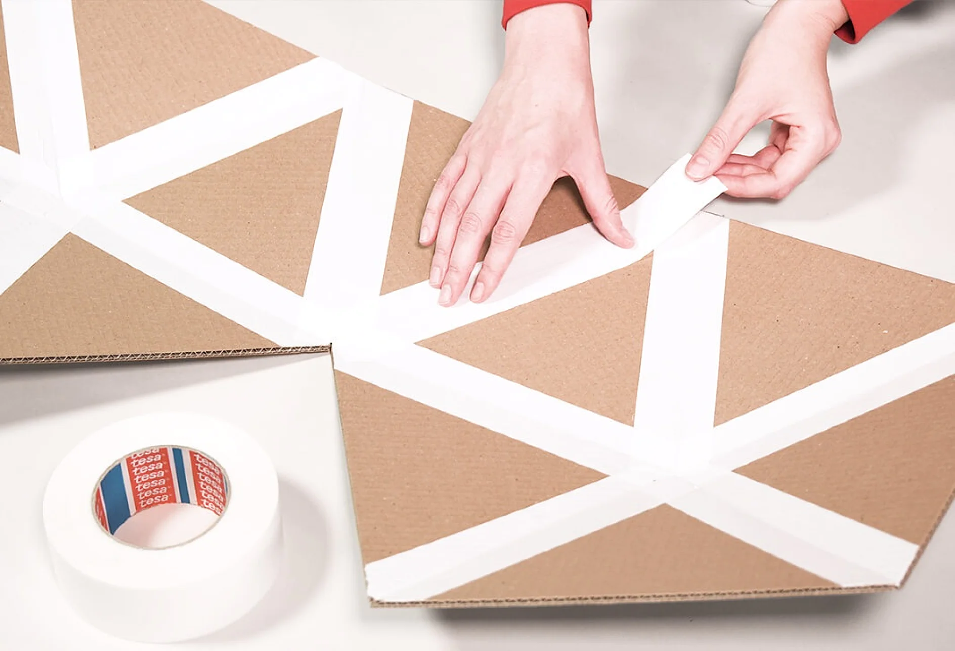 O persoană aplică bandă tesa albă pe un model geometric desenat pe o bucată de carton maro. O rolă de bandă tesa similară se află în apropiere, pe o suprafață deschisă la culoare. (Acest text a fost generat de inteligența artificială)