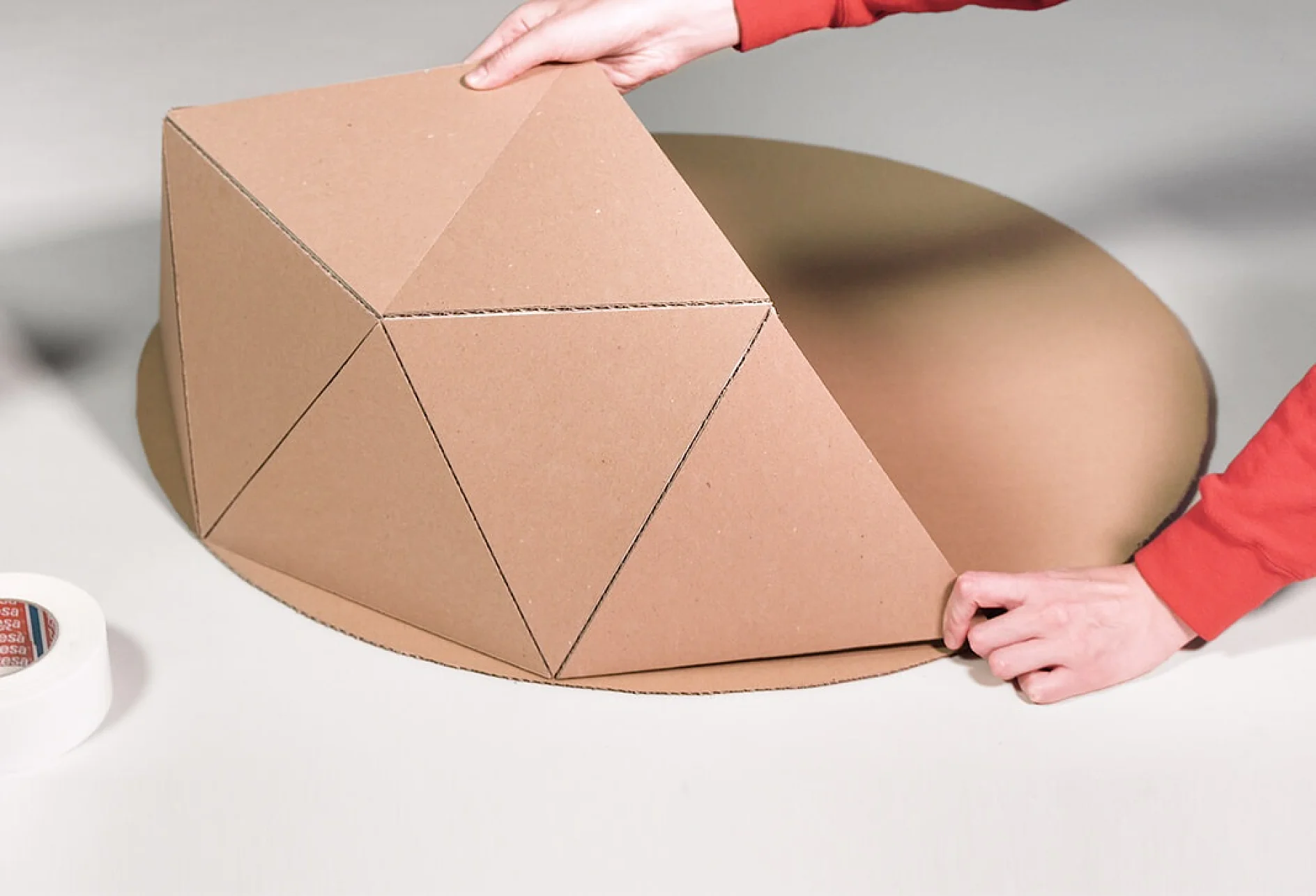 O persoană îmbrăcată cu un top roșu asamblează o structură geometrică din carton pe o bază rotundă. Structura este formată din secțiuni triunghiulare care sunt pliate în sus. În stânga este vizibil un sul de bandă adezivă tesa. (Acest text a fost generat de inteligența artificială)