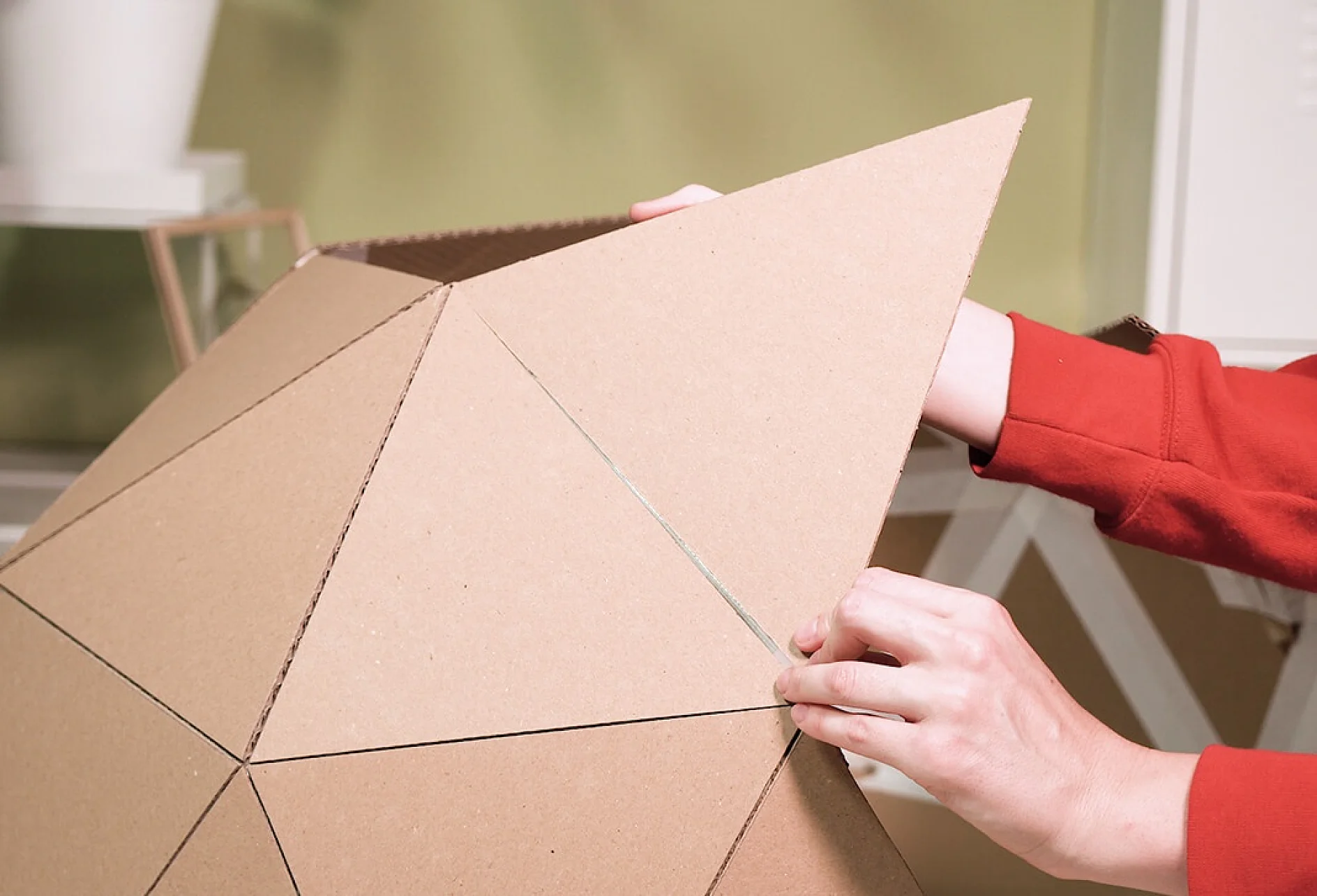 O persoană într-un pulover roșu asamblează o structură geometrică formată din piese triunghiulare de carton. Mâinile sale ajustează unghiurile formei folosind bandă adezivă tesa. Fundalul este un interior de casă neclar, cu un perete verde și mobilier alb. (Acest text a fost generat de inteligența artificială)