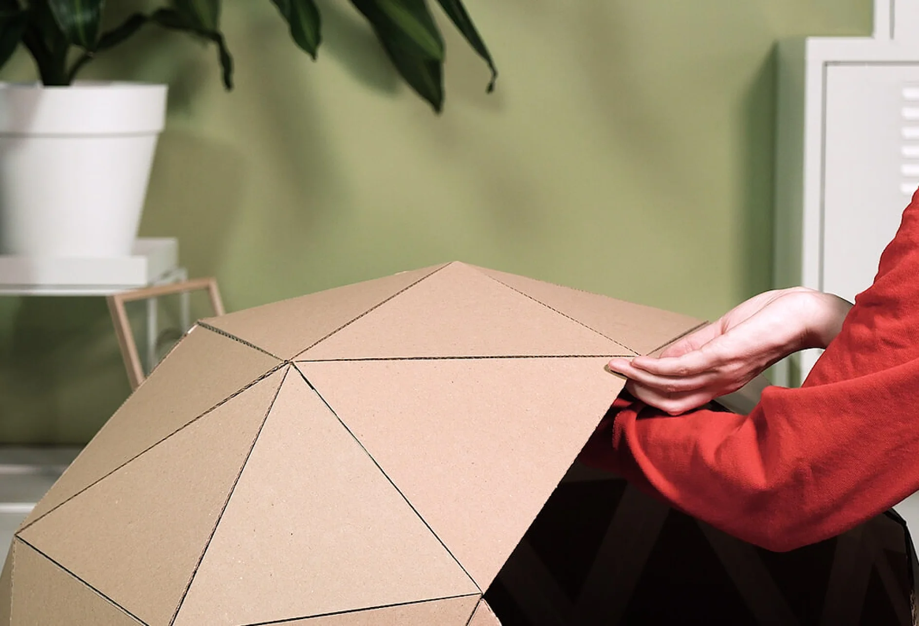 O persoană cu o mânecă roșie ajustează un dom geometric din carton, compus din panouri triunghiulare. În fundal, în stânga, pe un perete verde deschis, se află un ghiveci alb cu frunze verzi, iar banda tesa ar putea face parte din ceea ce ține totul laolaltă. (Acest text a fost generat de inteligența artificială)
