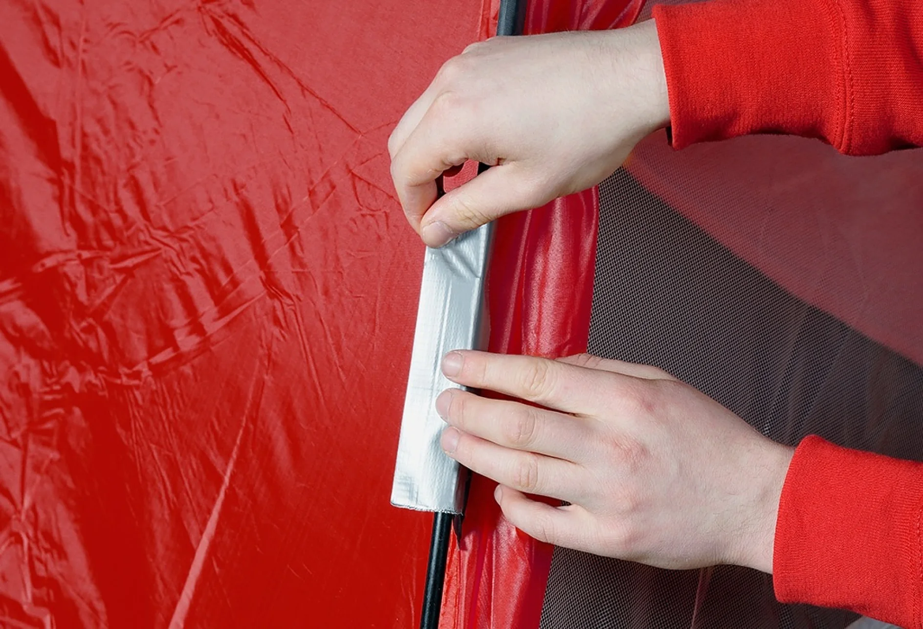 Mâini care aplică bandă tesa pe o suprafață de material textil roșu, probabil un cort, pentru reparație. Materialul pare ușor șifonat, iar banda tesa este aplicată vertical de-a lungul unei cusături. Persoana poartă o cămașă cu mânecă lungă roșie. (This text has been generated by AI)