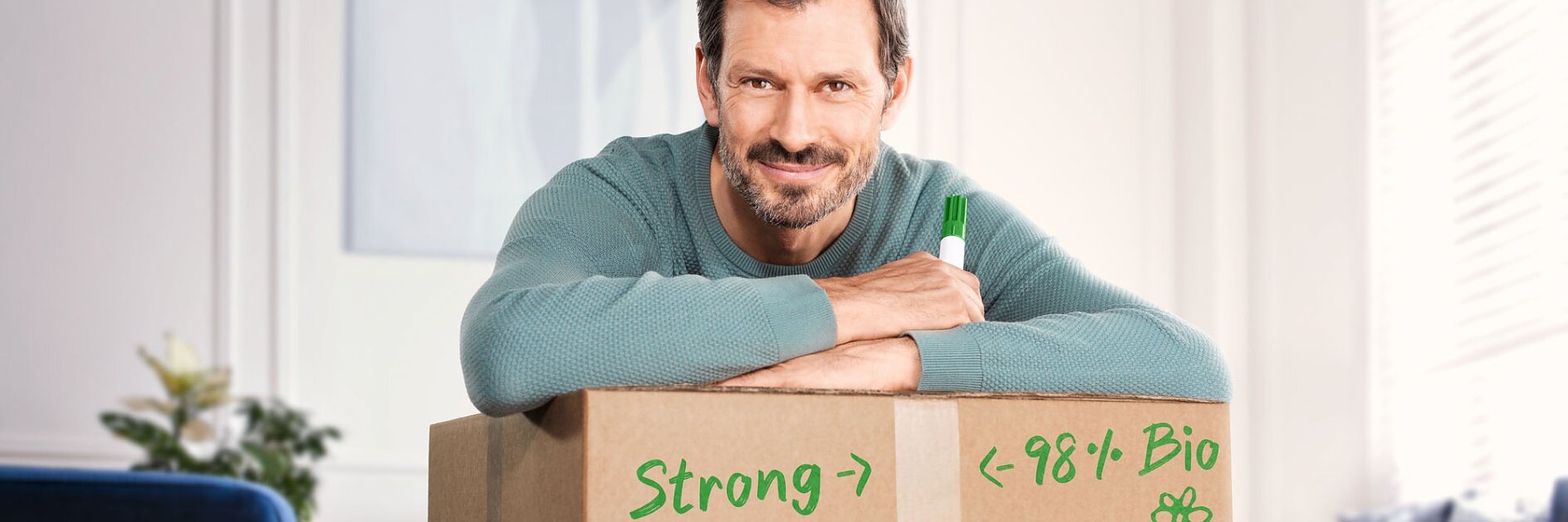 A man with short hair and a beard leans on a cardboard box, smiling. He is wearing a light blue sweater and holding a green marker. The box is labeled with hand-drawn text, “Strong” and “98% Bio” accompanied by a small flower sketch. The edges of the box appear to be secured with strips of tesa tape, ensuring everything inside stays safe and protected. (This text has been generated by AI)