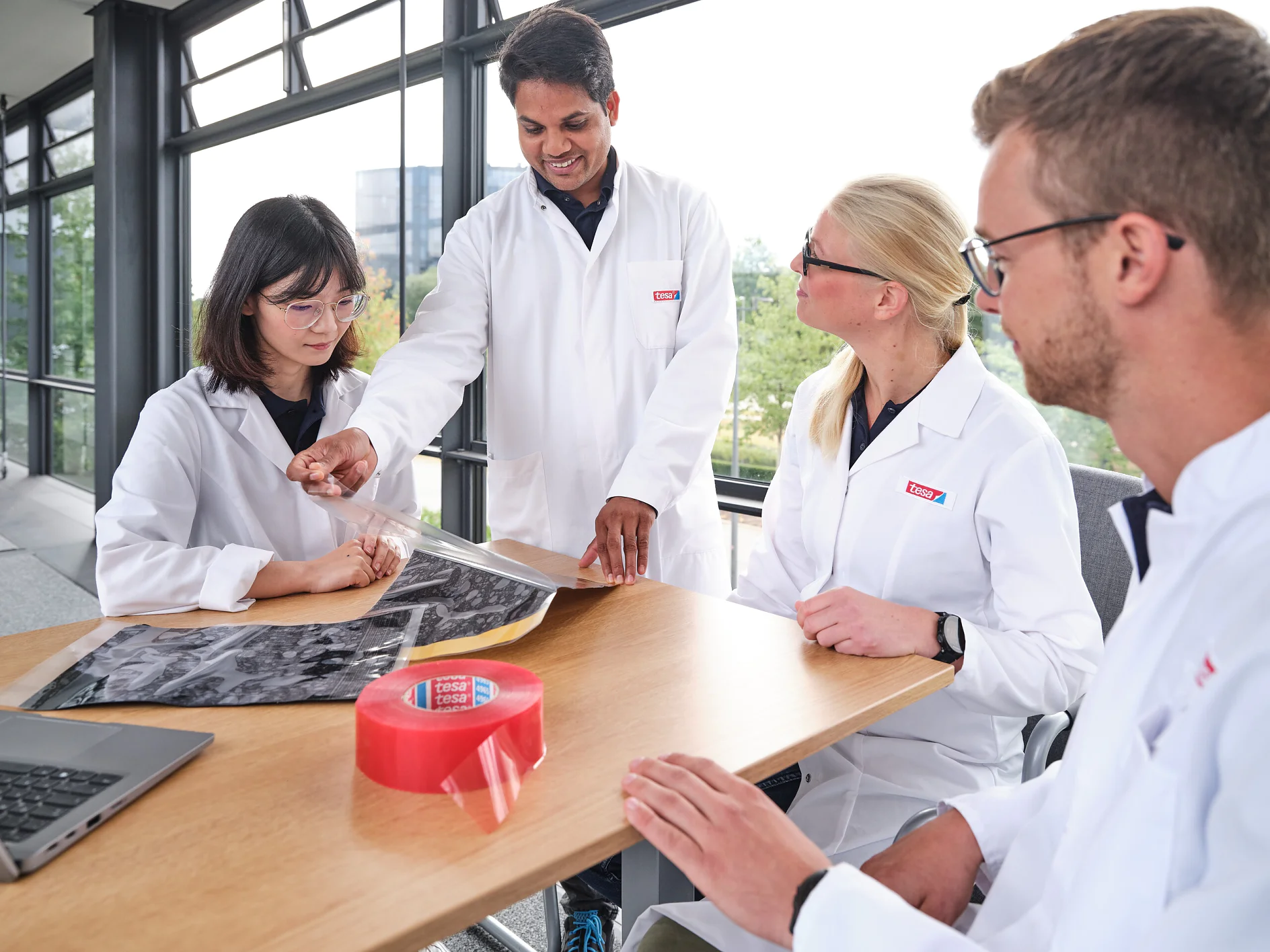 Four people in lab coats are gathered around a wooden table in a bright room with large windows. One person is showing a large sheet of material to the others. There is a roll of red tesa tape and a laptop on the table. (Acest text a fost generat de inteligența artificială)