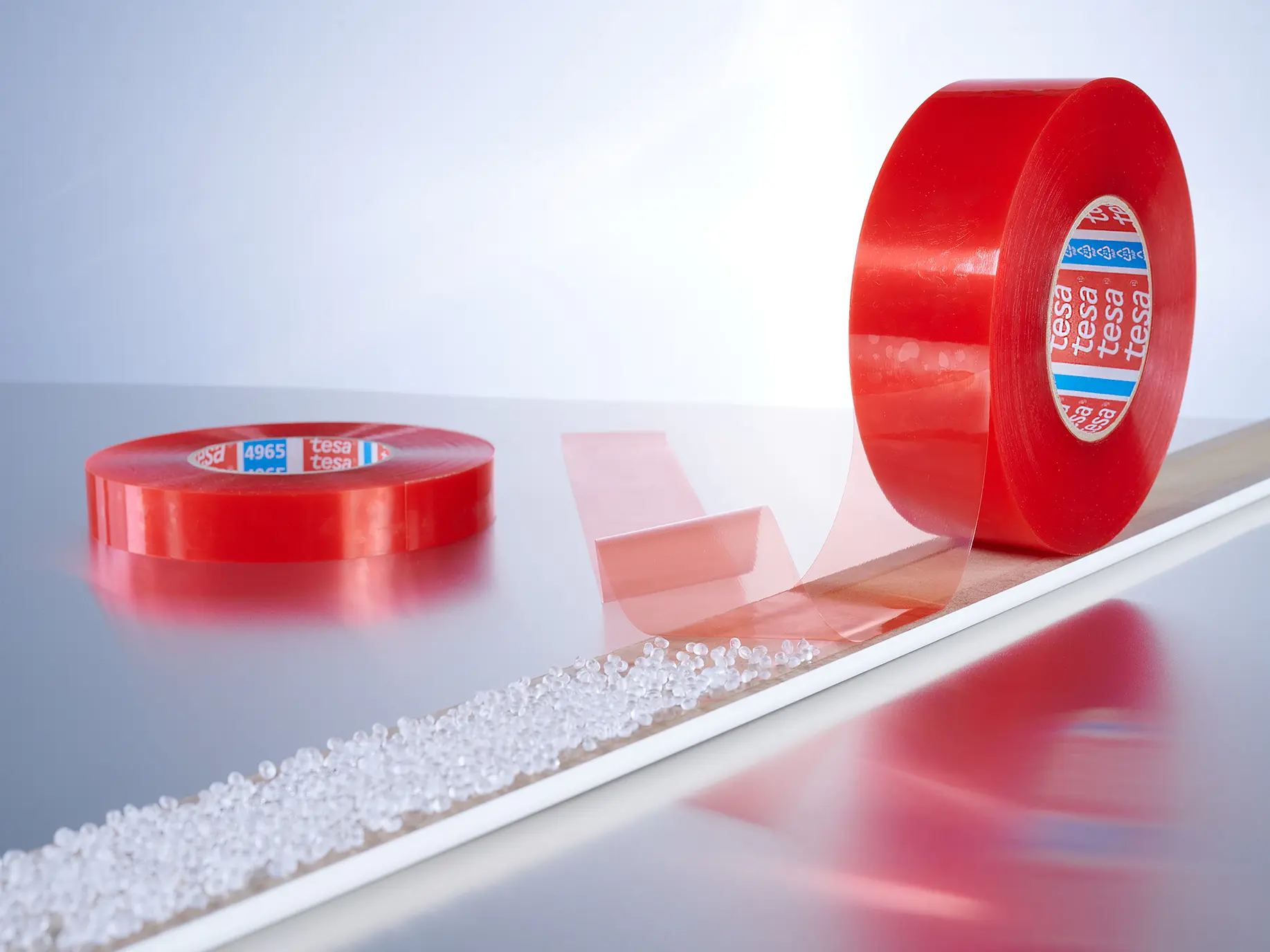 Two rolls of red adhesive tesa tape are displayed against a light background. One roll is lying flat, and the other is standing upright with a strip of tesa tape partially unrolled and stretched out along a line of small white granules.