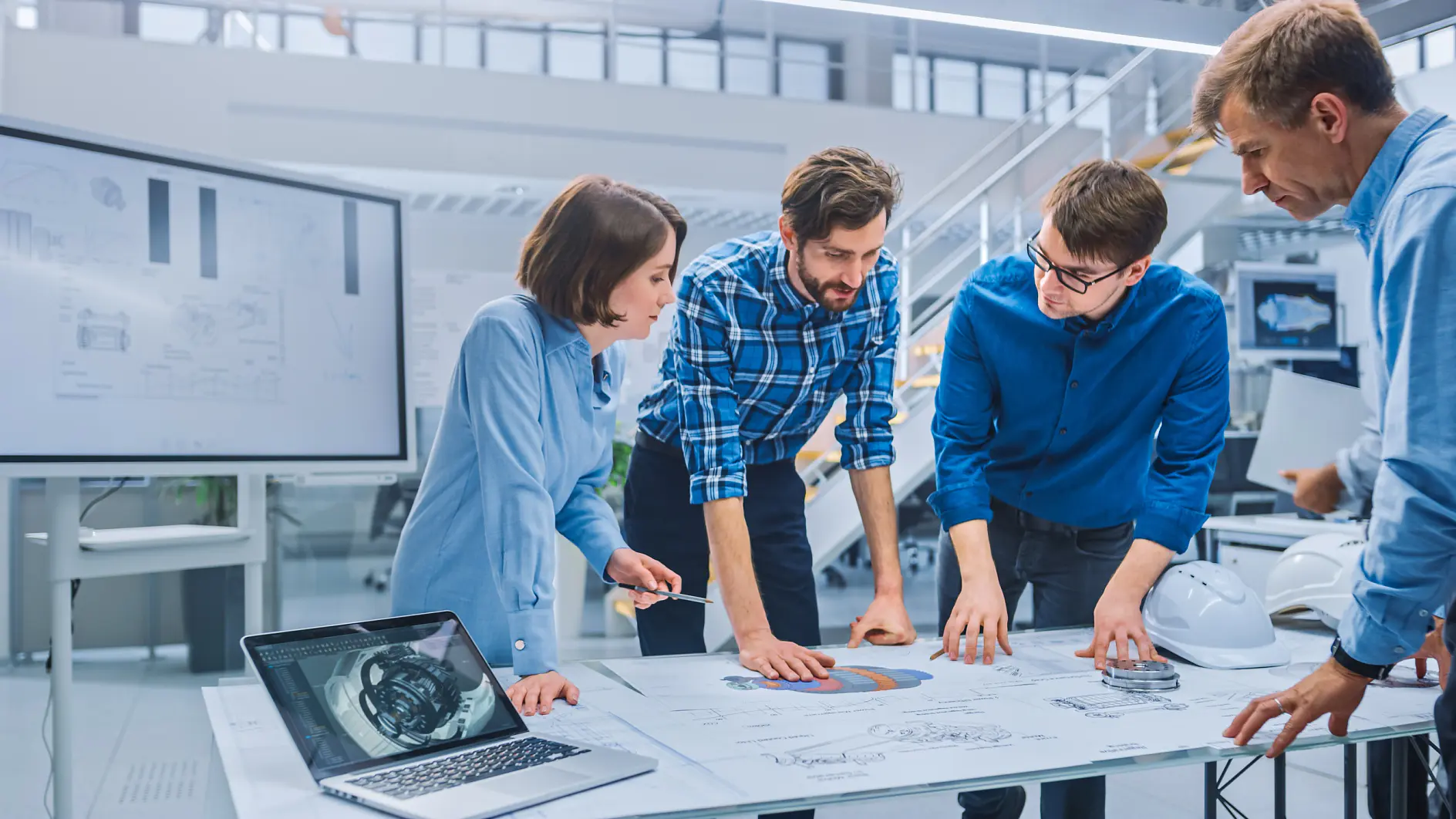 Four people are gathered around a table covered with blueprints and technical drawings in a modern office. They are engaged in discussion, and one person points at the plans. A laptop displaying a mechanical design, a large screen, helmets, and several rolls of tesa tape are visible. (Acest text a fost generat de inteligența artificială)