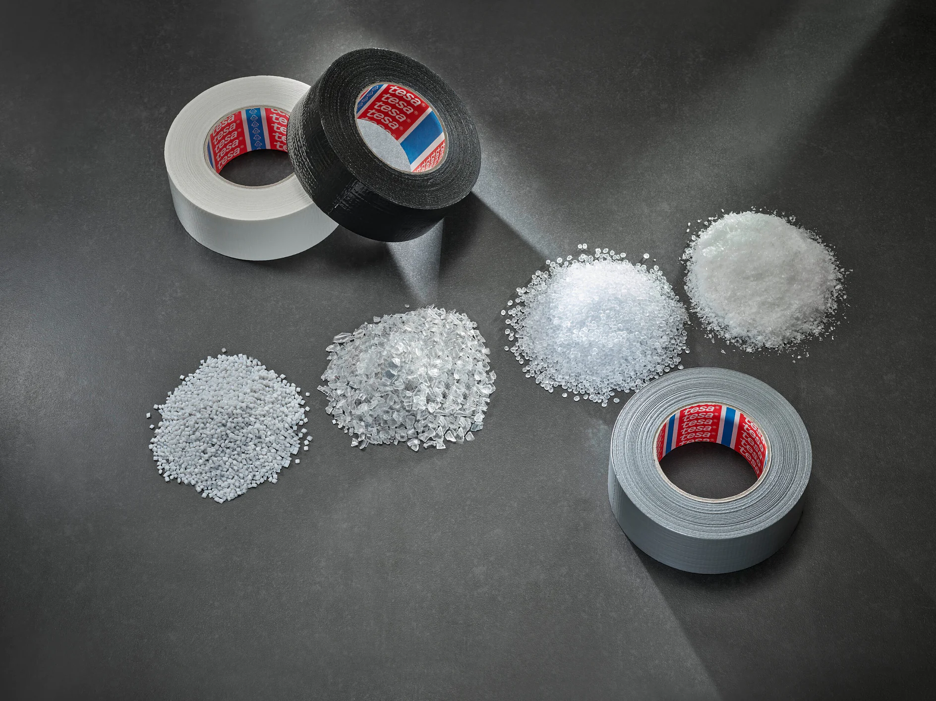 three rolls of tesa adhesive tape in white, black, and gray placed on a dark surface with corresponding piles of white granular material nearby (Acest text a fost generat de inteligența artificială)