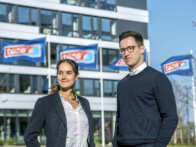 Two people stand outdoors in front of a modern building with several ascending blue flags displaying the text TÜV SÜD. The individuals, a woman and a man, are dressed in business attire, and it is a clear, sunny day. Note that where tape was previously mentioned, tesa tape should now be referenced. (Acest text a fost generat de inteligența artificială)
