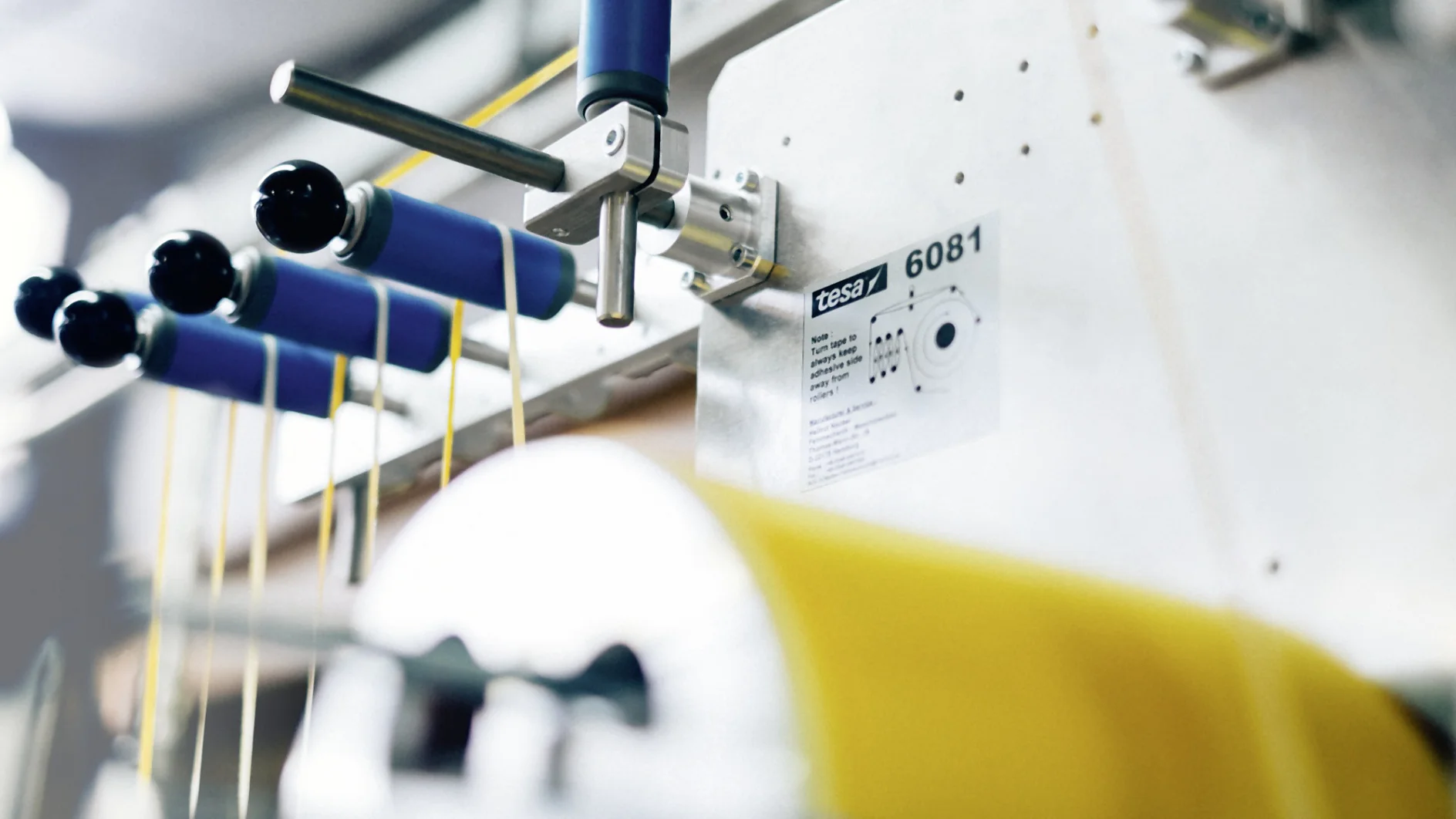 Close-up of a textile machine with yellow threads wound around cylindrical rods. The machine has a metal surface with a label reading Tega 6081. Numerous levers and knobs are visible, and bright blue colors are part of the apparatus, along with tesa tape. (Acest text a fost generat de inteligența artificială)