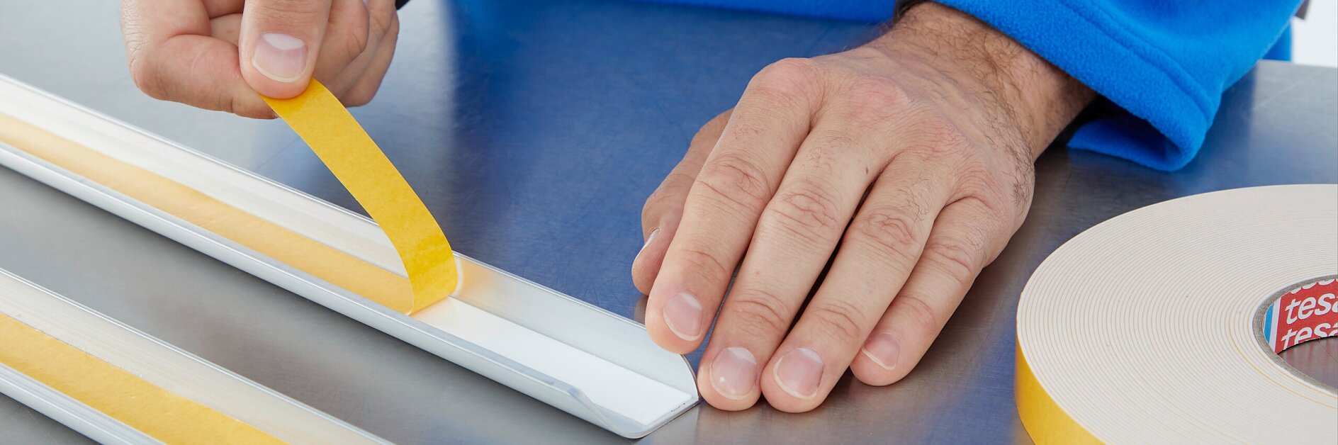 A person in a blue sleeve applies a strip of yellow double-sided tesa tape to a white rectangular object on a metallic surface. A roll of similar tesa tape is visible on the right side of the image. (Acest text a fost generat de inteligența artificială)