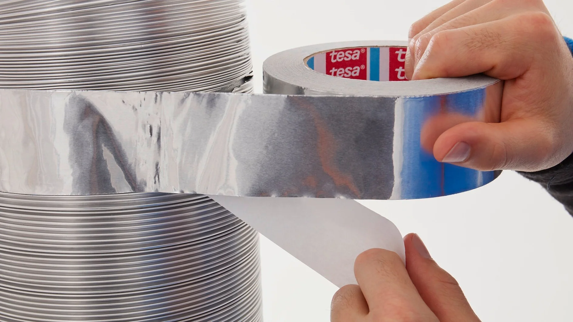 A person is applying a strip of metallic tesa tape onto a cylindrical object with horizontal ridges. The tesa tape is being unwound from a roll held in one hand, while the other hand removes the backing paper. (Acest text a fost generat de inteligența artificială)