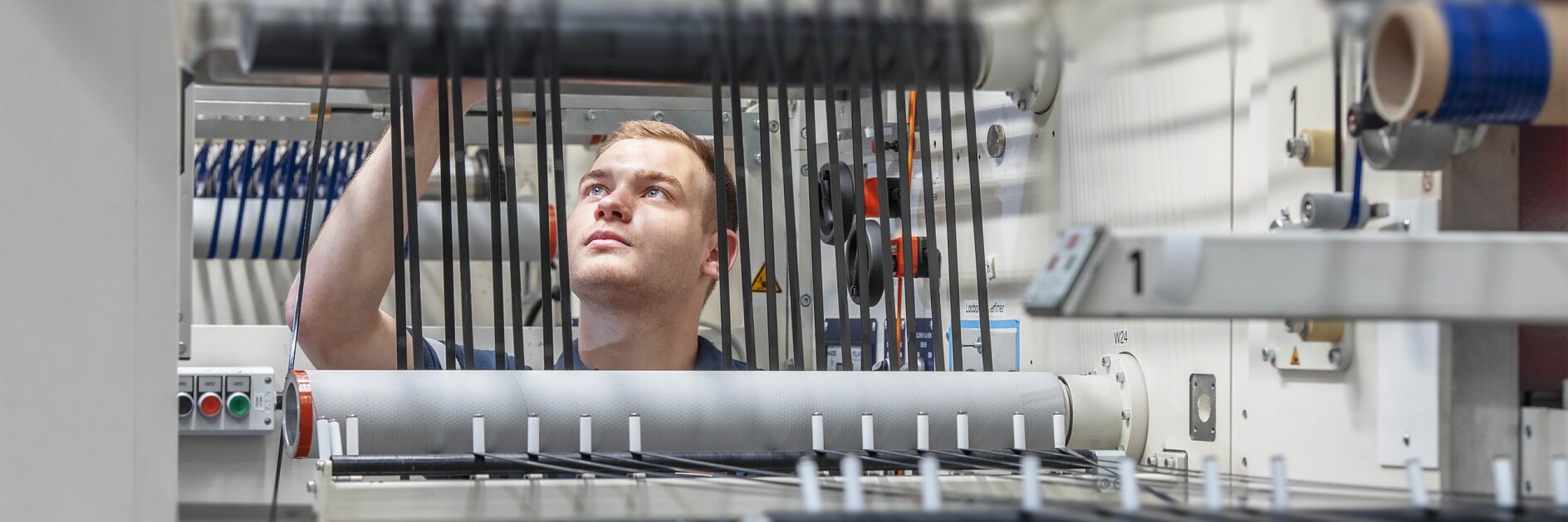 A person in a blue uniform works with machinery in a factory setting, adjusting black cables on a series of rollers and levers using tesa tape. The environment is industrial, with various mechanical components and controls visible. (Acest text a fost generat de inteligența artificială)
