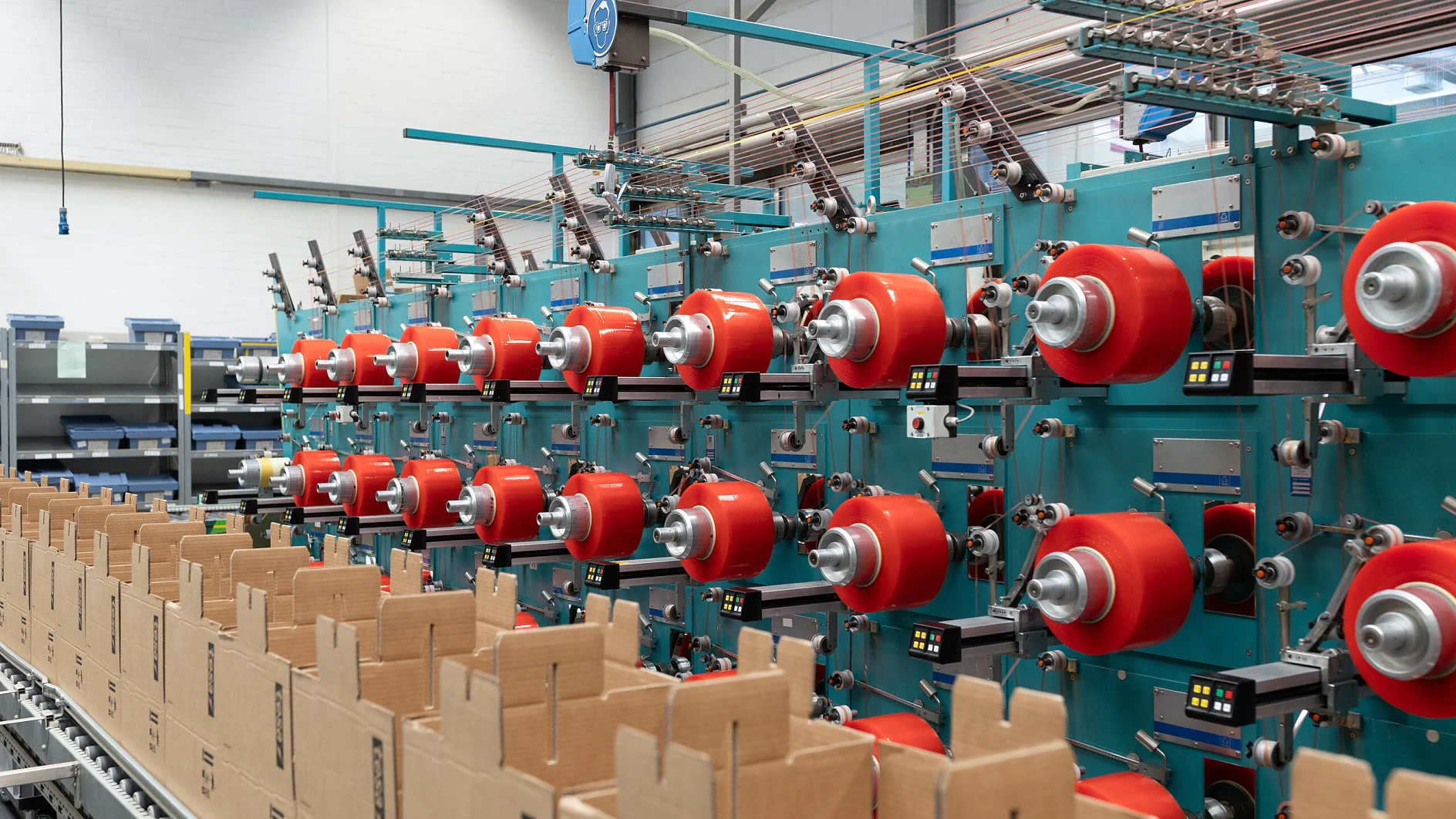 A factory interior showing a row of teal-colored machines with multiple red spools of thread and rolls of tesa tape. Cardboard boxes are aligned on a conveyor belt in the foreground. The setting appears organized, with equipment and materials neatly arranged. (Acest text a fost generat de inteligența artificială)