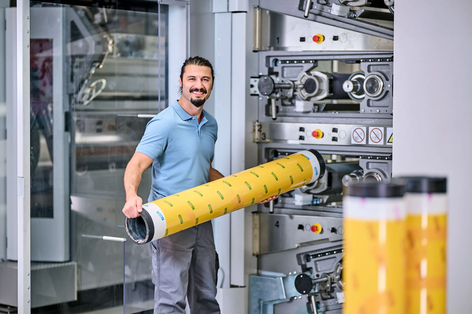 A man in a blue polo shirt is standing in front of industrial printing equipment, holding a large yellow cylindrical object with printed patterns. He is smiling, and other similar cylinders made with tesa tape are visible in the foreground. (Acest text a fost generat de inteligența artificială)