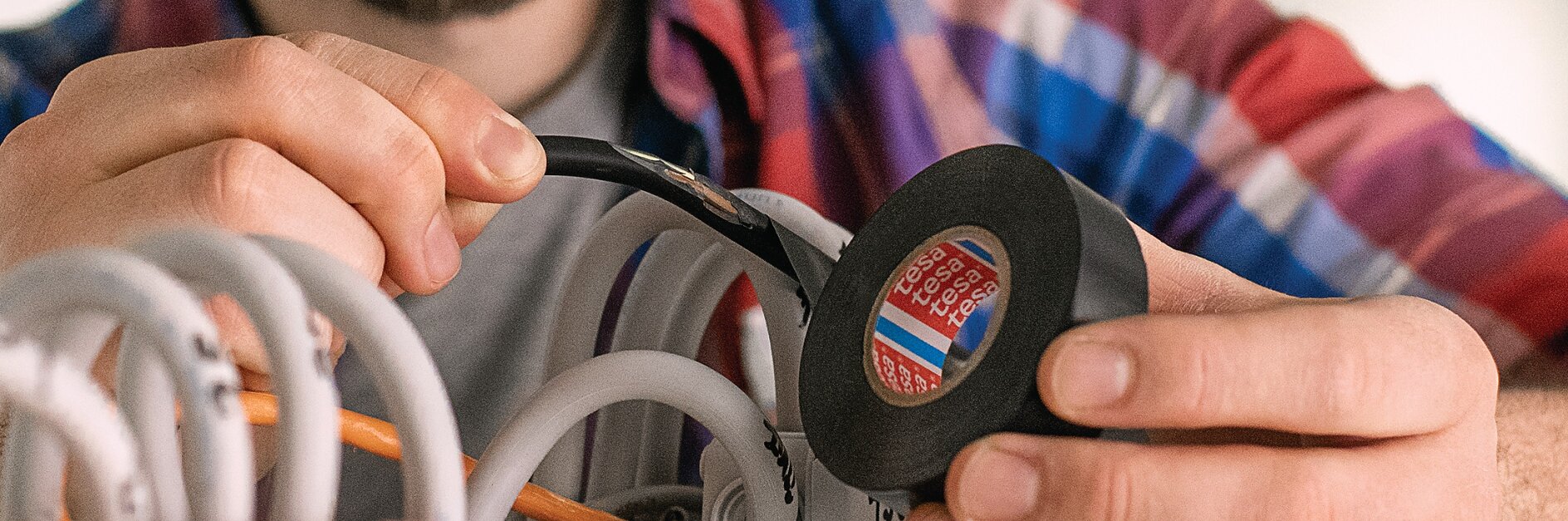 A person wearing a plaid shirt is holding a roll of black tesa tape and is applying it to wires in an electrical box. The scene appears to involve electrical work or installation, with multiple cables being organized. (Acest text a fost generat de inteligența artificială)