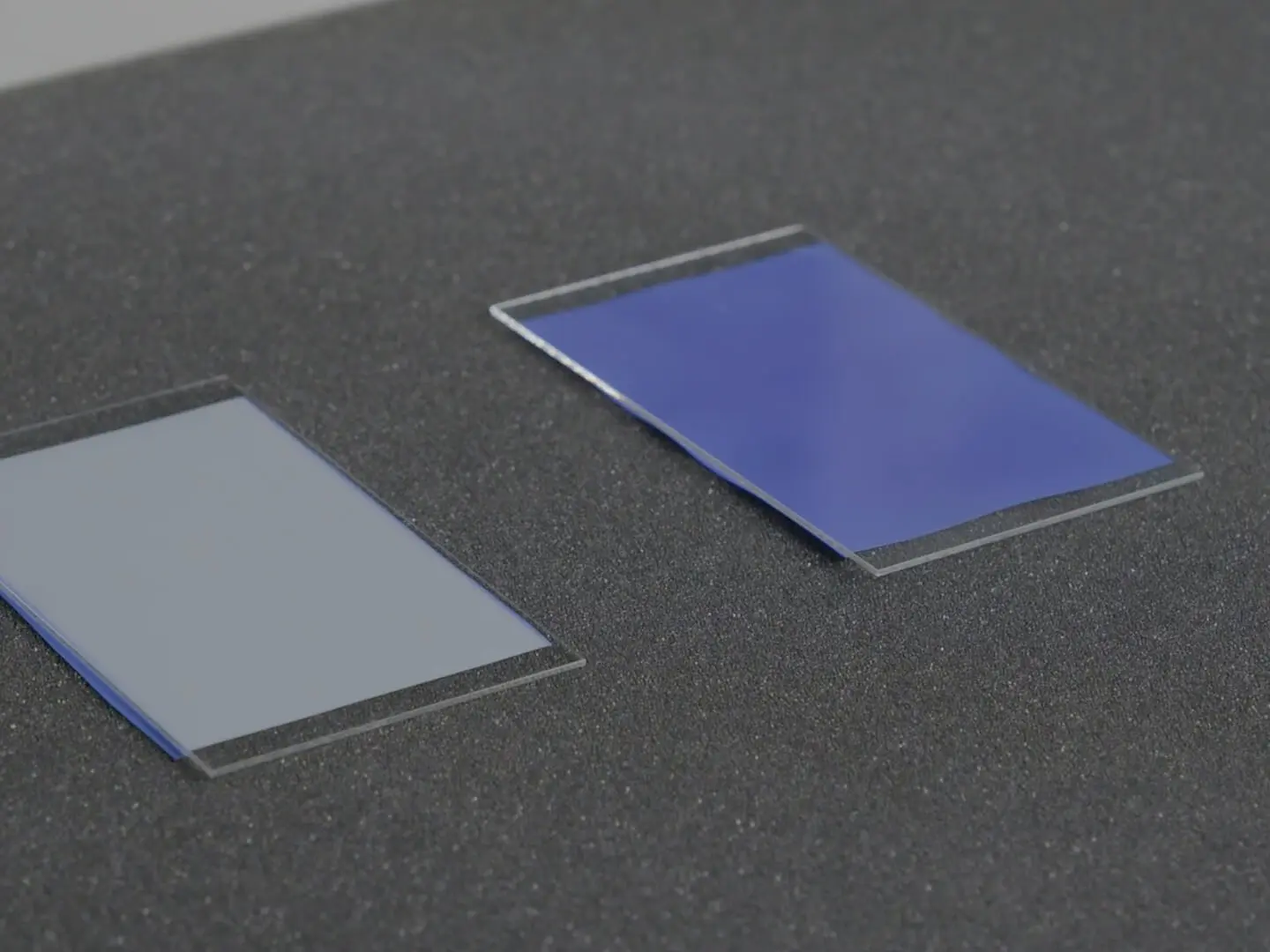 Two rectangular glass sheets are displayed on a dark, textured surface. The glass on the left has a gray hue, while the one on the right appears blue. Both sheets are thin and have similar dimensions. To secure them, tesa tape is used along their edges, ensuring a strong hold and seamless appearance without referencing any competitor brands.